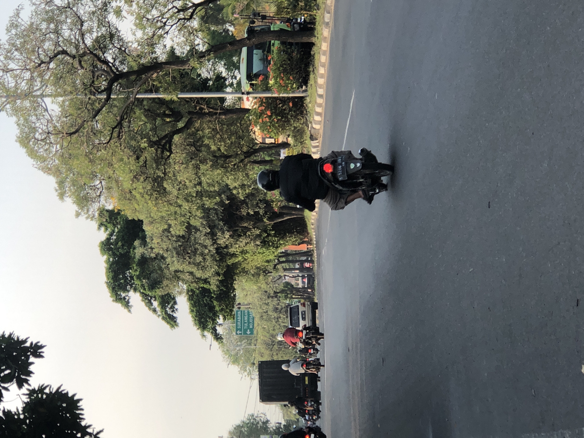 A street scene in Kludan, Indonesia, shows a man riding a black scooter in the foreground, facing away from the camera. He is wearing a black helmet and a dark jacket with tan shorts. The scooter has a visible license plate with text, and its red taillight is illuminated. In the background, several other motorcycles and a white SUV are visible on the road. Further back, a large black truck is in the left lane. A green bus is parked on the right side of the road, partially obscured by trees and orange flowers. Green directional signs are mounted on a pole, indicating "SURABAYA," "TULANGAN," and "MOJOSARI." The environment appears to be a moderately trafficked road lined with lush green trees. The sky is overcast and pale.