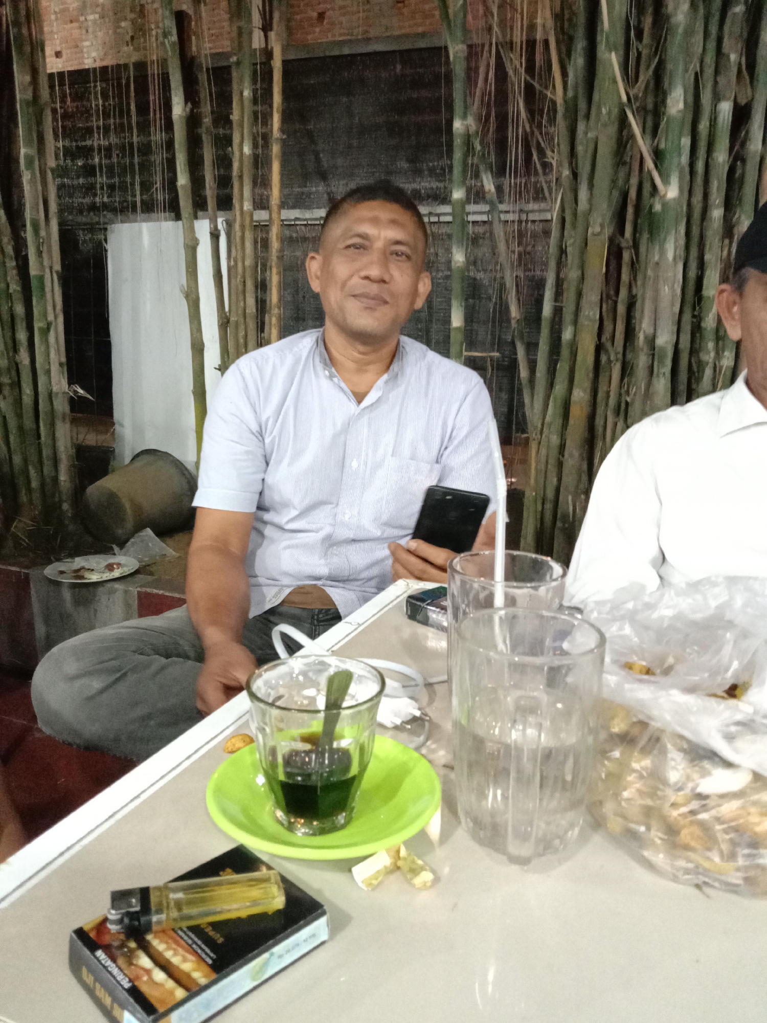 The image shows a man sitting at a table outdoors in Lhokseumawe, Indonesia. He is holding a smartphone and wearing a light blue, button-down shirt and gray jeans. Another man is partially visible to his right, wearing a white shirt and a black cap.
On the table are several items: a small glass of dark green liquid on a green saucer, a tall glass of water with a straw, a plastic bag filled with snacks, a pack of cigarettes, and a lighter. The background consists of a brick wall, white wall, and bamboo stalks. A pot sits on a ledge behind the seated man.
