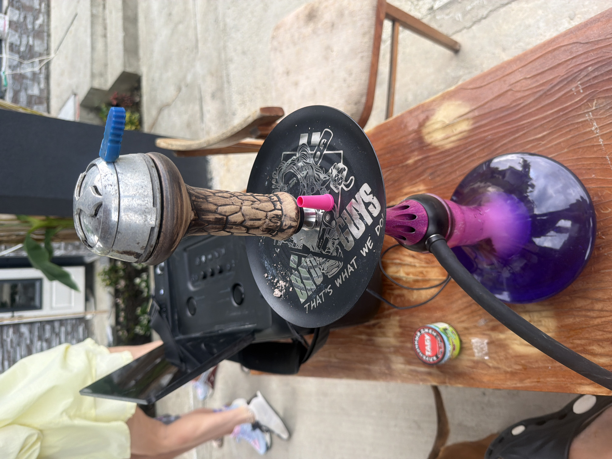 This media file depicts a close-up, rotated view of a hookah setup on a wooden table. The main subjects are the hookah components: a metallic base with intricate carvings, a wooden stem, a black disc with white graphics, a purple hose, and a violet glass base containing a purple liquid. A small, blue, ridged piece, likely a poker or valve, is visible on top of the metallic base. A small, pink object, possibly a grommet, is attached to the stem. A small, circular tin, possibly containing tobacco or accessories, is also on the table.
The scene appears to be outdoors, with hints of a paved ground and possibly a building or wall in the background. A chair with a wooden frame and what appears to be a fabric seat is partially visible behind the hookah. The lighting suggests it might be daytime, with some shadows indicating natural light. There are no people prominently featured in the shot, though a sliver of a person's arm and legs, wearing light-colored clothing and white sneakers, is visible at the bottom left, suggesting someone is either setting up or using the hookah.
The black disc on the hookah stem features graphics and text. The text visible reads "UNS THAT'S WHAT WE DO". The overall impression is a relaxed, informal setting, possibly a backyard or patio, where someone is preparing to use the hookah. The location is identified as Baranovka, Russia.