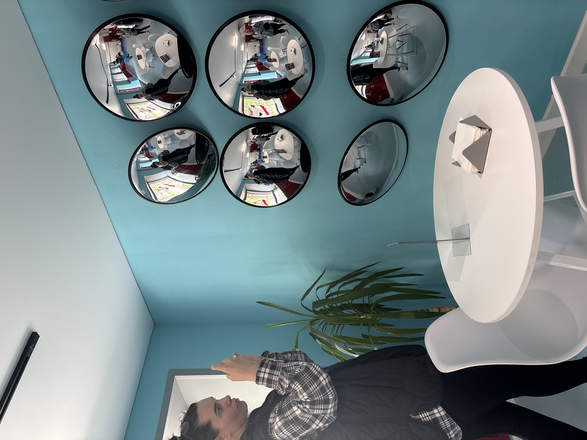 In the city of Chaikovsky, Russia, a woman is seen from the side, wearing a black vest over a plaid shirt and black leggings, as she holds up a smartphone and takes a picture. She stands near a tall, green potted plant. To her right, a wall is decorated with multiple convex mirrors, reflecting a bright, modern interior. These mirrors display distorted images of people, white tables, and chairs within what appears to be a cafe or communal space. The wall is painted a vibrant turquoise. In the foreground, a white round table with a gray napkin holder is visible, with white chairs around it. A light fixture is mounted on the ceiling.