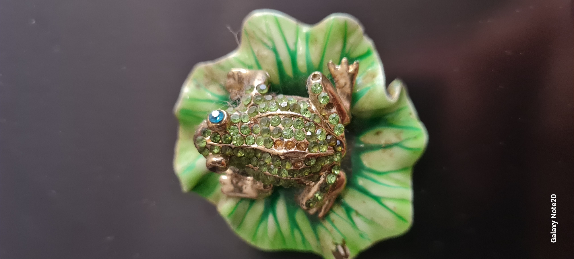 A close-up, high-angle shot shows a small, ornate, gold-colored frog figurine encrusted with small, green rhinestones, with one blue rhinestone for its eye. The frog is perched on a green, leaf-shaped dish, also adorned with green enamel markings. The background is a dark, blurred surface. The image was taken in Holon, Israel, as indicated by the "Galaxy Note20" watermark at the bottom left, which is a phone model commonly used in Israel, and Holon's known presence of digital infrastructure supporting such devices.