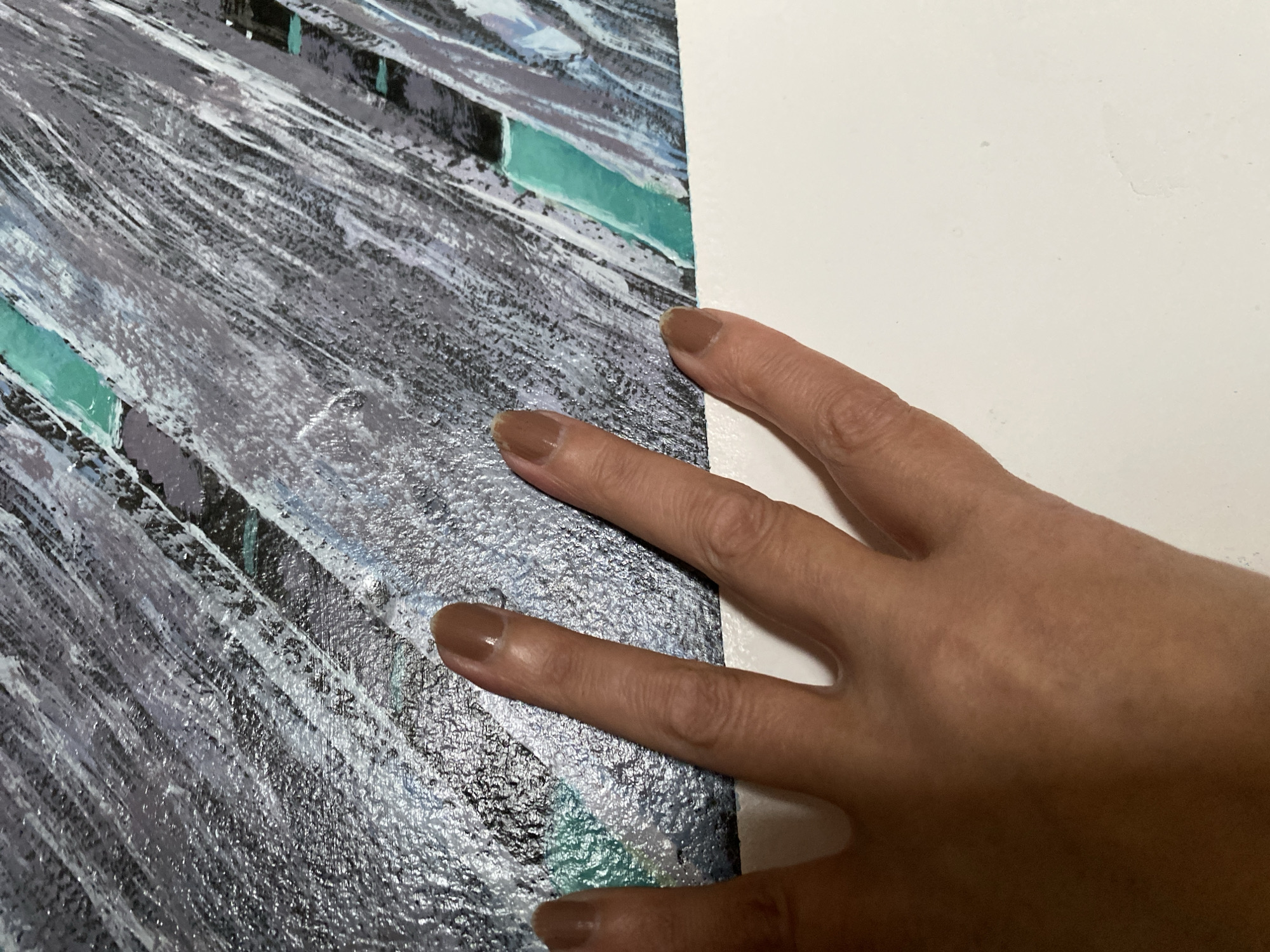 The image shows a close-up of a hand with light brown skin and light brown nail polish, resting on a textured surface with streaks of gray, black, white, and teal. The hand is positioned in the lower left and center of the frame, with four fingers and a portion of the thumb visible. The fingers are extended and gently pressed against the textured surface, which appears to be a painting or a textured wallpaper. The background is a stark white surface, likely a wall or a table, occupying the bottom portion of the image. The image is taken from a slightly elevated angle, looking down at the hand and the textured surface. There is no indication of the specific location within Bílina, Czechia.