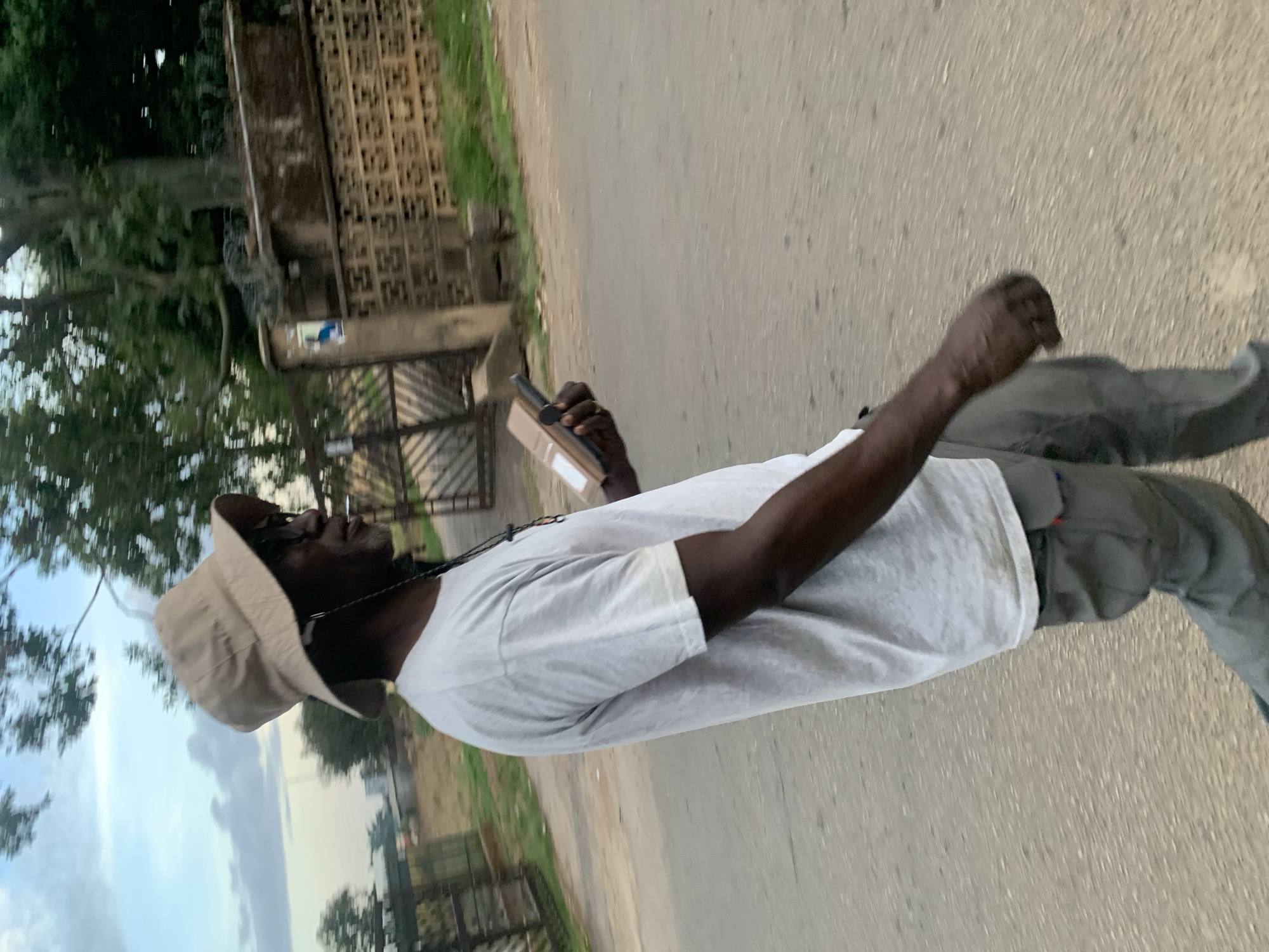 An adult Black man is walking on an asphalt road with a phone in his hand. He is wearing a light-colored t-shirt, grey pants, and a light-colored bucket hat. A cigarette is in his mouth. A building and some trees are visible in the background. The location is likely Jos, Nigeria, based on visual cues and the prompt.