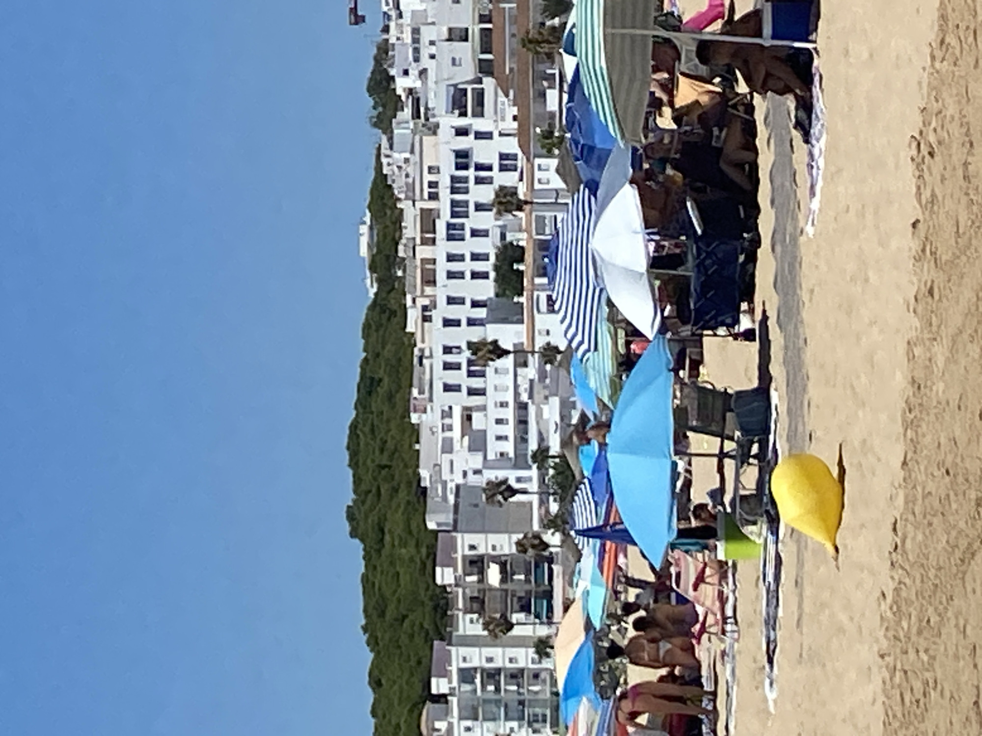 The image depicts a crowded beach scene in Conil de la Frontera, Spain. In the foreground, a yellow inflatable object lies on the sand. The midground is filled with numerous beach umbrellas providing shade for beachgoers, some of whom are seated on chairs or towels. Beach tents and trolleys are also visible. In the background, white buildings with multiple stories are clustered together, backed by a dense green hillside. The sky is clear and blue.