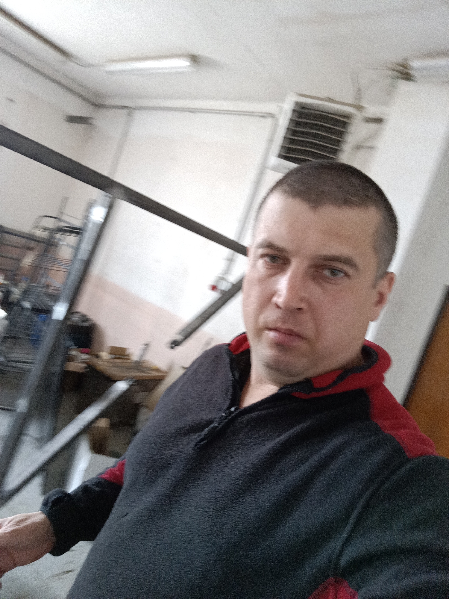 A man takes a selfie in what appears to be a workshop or industrial setting. The man has short, dark hair and is wearing a black and red fleece jacket. He is looking directly at the camera. In the background, there is industrial equipment, including metal beams and shelving, as well as a fluorescent light fixture and an air conditioning unit on the wall. The overall environment suggests a workspace.