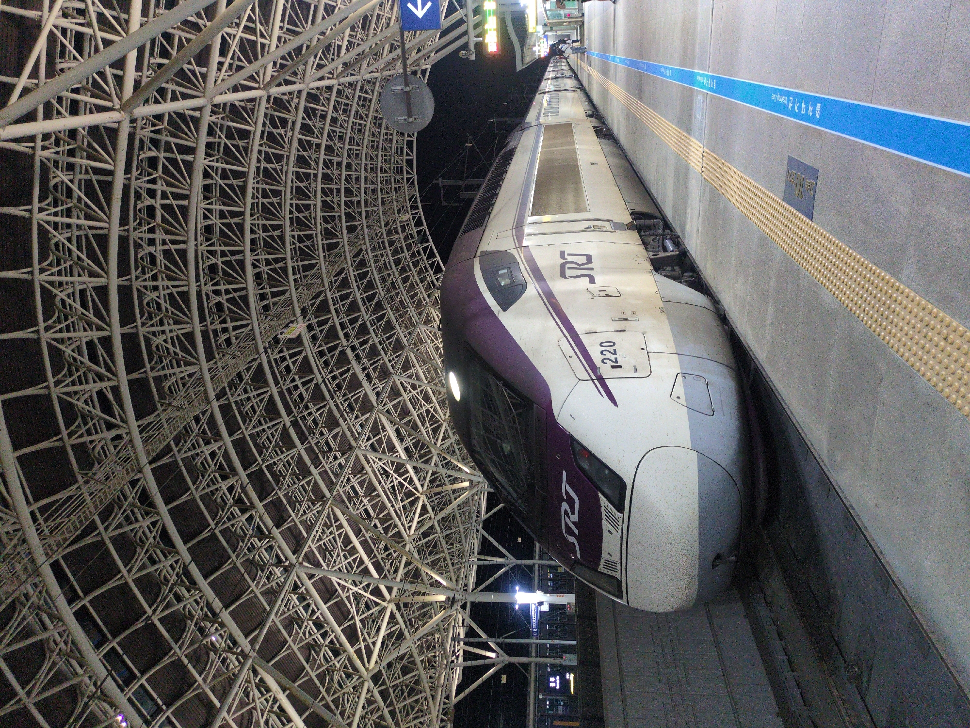 A high-speed train is stopped at the platform of Singyeongju Station in Gyeongju-si, South Korea. The train, predominantly white with a purple stripe, is labeled with the "SRT" logo and the number "1220." The station platform, marked with a blue line and textured warning surfaces, is adjacent to the train. Above the train, a network of white, crisscrossing metal beams forms a canopy over the station. A blue sign with a white arrow hangs from the structure. In the background, a night sky and distant city lights are visible.