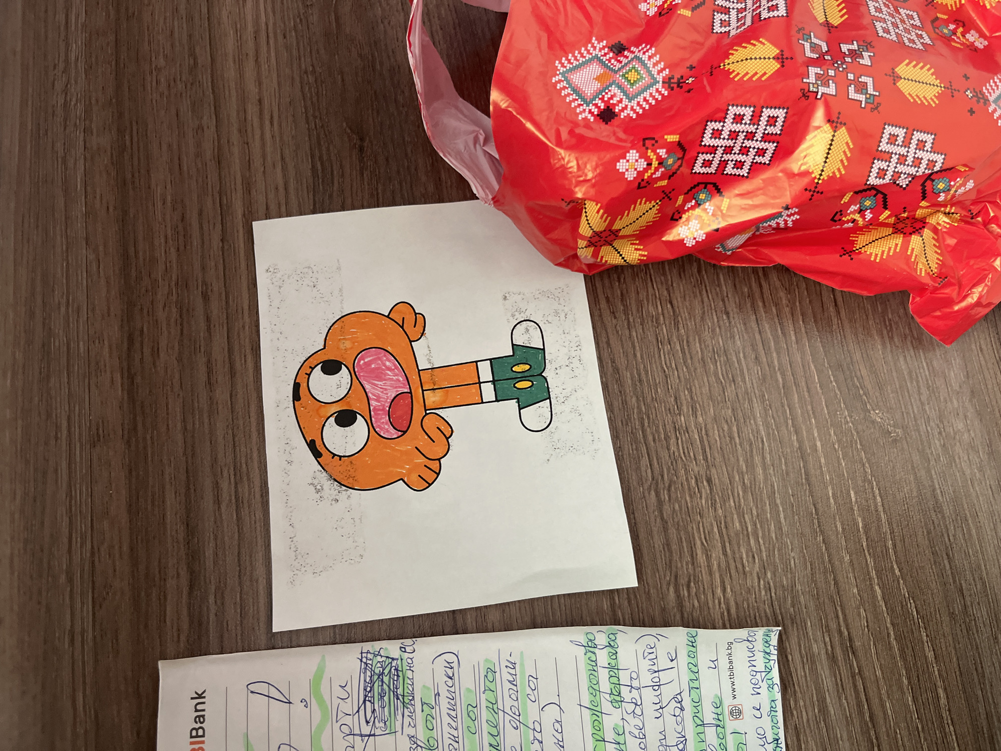 A bright red, crinkled plastic bag with a traditional Bulgarian geometric pattern lies on a brown wooden table. The bag is partially open, revealing a glimpse of a pink interior. Next to the bag is a piece of white paper featuring a colorful drawing of a cartoon character. The character is orange with large, surprised eyes and a wide mouth. It has black pupils, pink cheeks, and is wearing green shorts and white socks with green stripes. The drawing is simple and childlike. Below the drawing, another piece of paper is visible, with handwritten text in blue and green ink. Part of the text includes the website "www.tblbank.bg". The scene appears to be indoors, with the wooden table suggesting a domestic setting. The lighting is even, indicating it is likely daytime. There are no people visible in the image. The overall impression is of a casual, possibly creative moment, with a child's drawing and a decorative bag. The location context is Vratsa, Bulgaria.