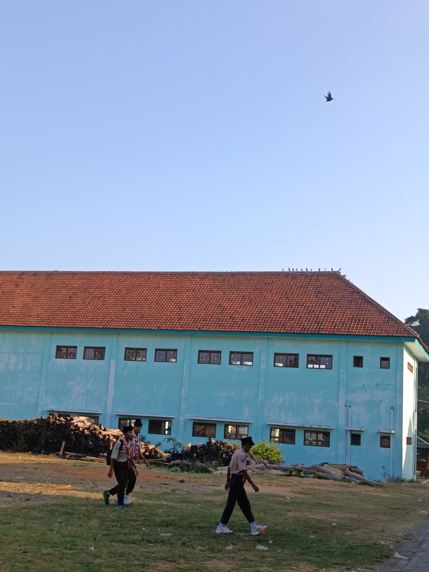 A long, light blue building with a reddish-brown tile roof dominates the mid-ground of the image. The building has numerous rectangular windows with dark frames. Several birds are perched on the edge of the roof. In the foreground, two individuals in scout uniforms are walking across a grassy area. One scout is in the foreground on the right, facing left, wearing a dark hat and dark pants. The other scout is further back and to the left, also facing left, wearing a similar uniform with a red and white neckerchief. Behind the scouts, a large pile of cut logs is visible. The sky is clear and blue. A single bird is in flight in the upper right portion of the sky. The image appears to be taken outdoors during daylight. The location is Ramban Wetan, Indonesia.