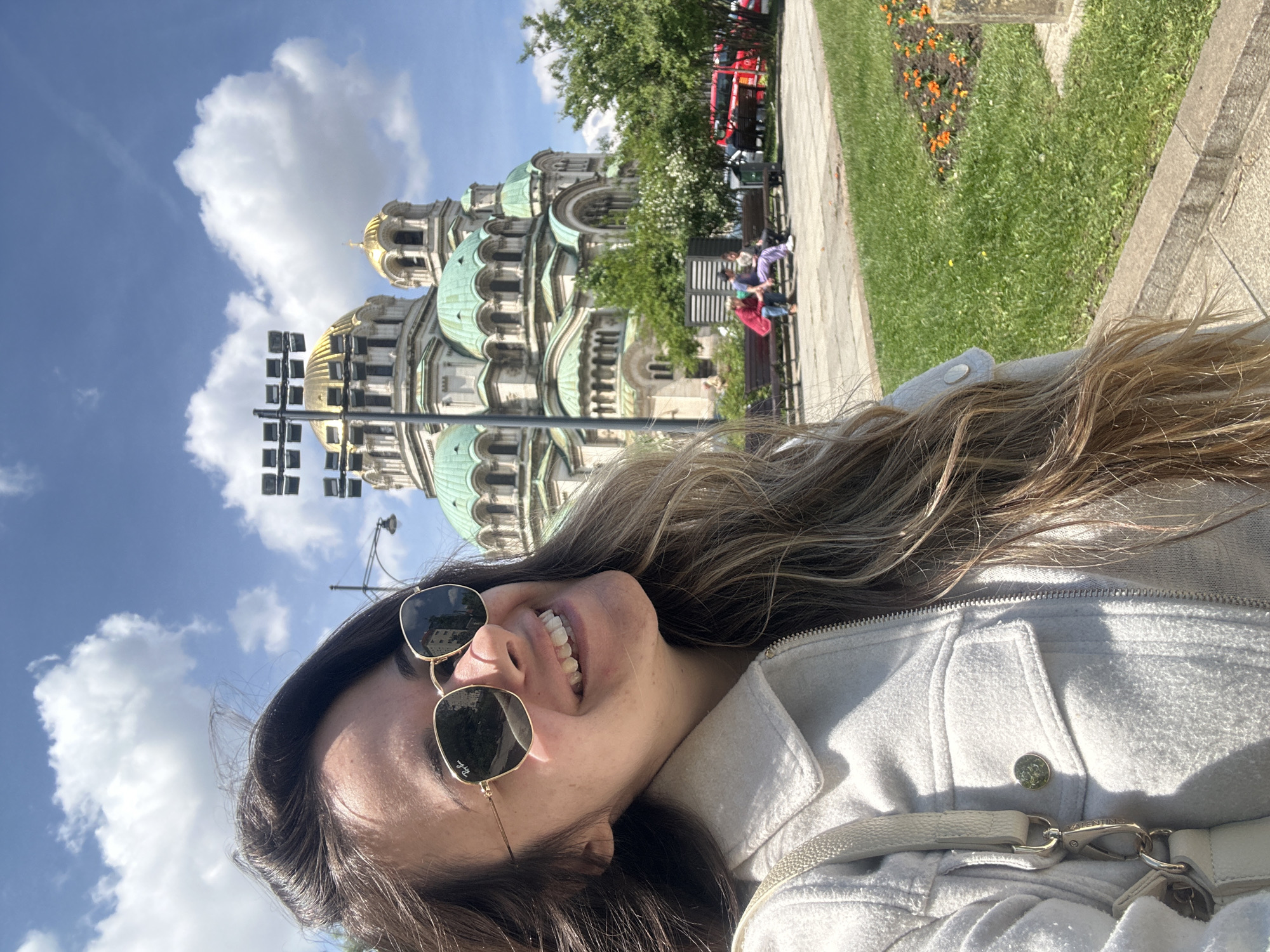 A woman with long brown hair and wearing sunglasses and a white jacket stands in front of the Alexander Nevsky Cathedral in Sofia, Bulgaria. She is smiling. The cathedral has green domes and golden tops. A streetlight stands to the left of the building. People sit on benches along a sidewalk next to grass and flowers. A red vehicle is parked behind trees. The sky is blue with fluffy white clouds.