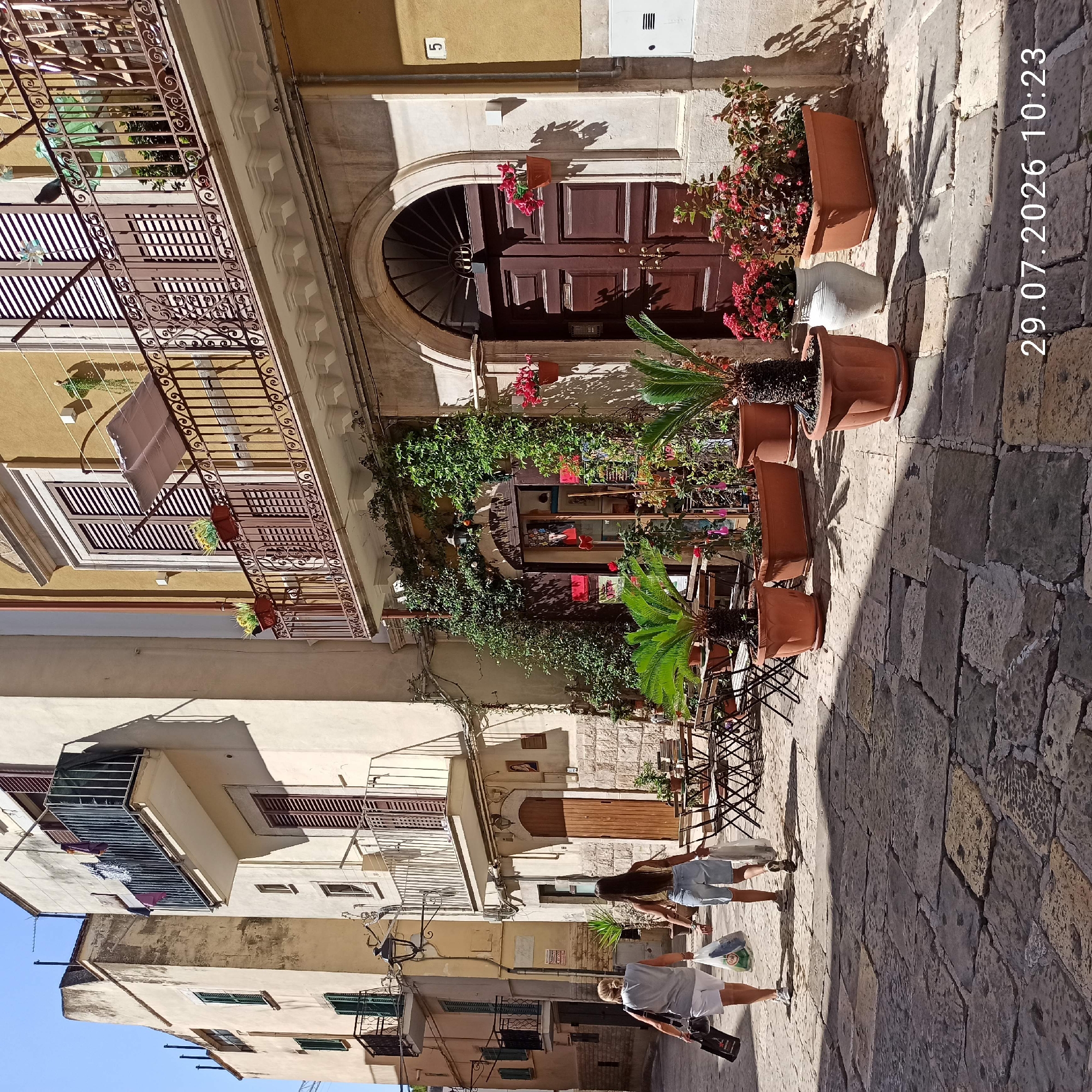 The image depicts a cobblestone street in Bitonto, Italy, lined with buildings featuring balconies and potted plants. Two women are walking away from the camera, each carrying shopping bags. One woman is wearing a grey t-shirt and white shorts, and the other is wearing a yellow tank top and denim shorts. In the foreground, there are several potted plants, including palm trees and flowering bushes, placed outside a building with a brown wooden door. A few tables and chairs are also visible on the street, suggesting an outdoor seating area. The buildings are a mix of cream and yellow colors, with traditional shutters on the windows. The scene is bathed in sunlight, casting shadows on the street and buildings. The date stamp in the bottom right corner reads "29.07.2026 10:23".