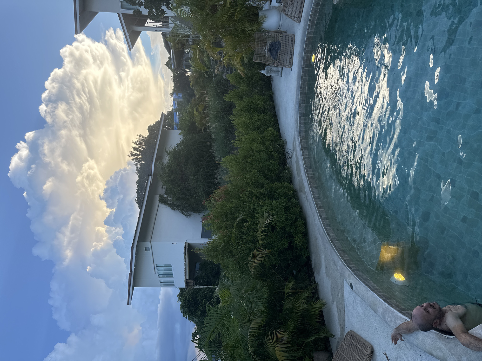 The image depicts an outdoor swimming pool area at dusk in Tambon Ko Pha Ngan, Thailand. In the foreground, a man is partially submerged in the pool, leaning on the edge. The pool water is a turquoise color and is rippled, reflecting the sky and surrounding lights. To the left of the pool, a light fixture is visible underwater, emitting a yellow glow. Behind the pool, lush green vegetation, including palm trees and bushes, surrounds the area. In the background, there are modern white buildings with angular roofs, characteristic of tropical architecture. The sky is dramatic with large, illuminated clouds against a darkening blue sky, suggesting the end of a day with possibly stormy weather. The scene conveys a sense of tropical relaxation and ambiance.