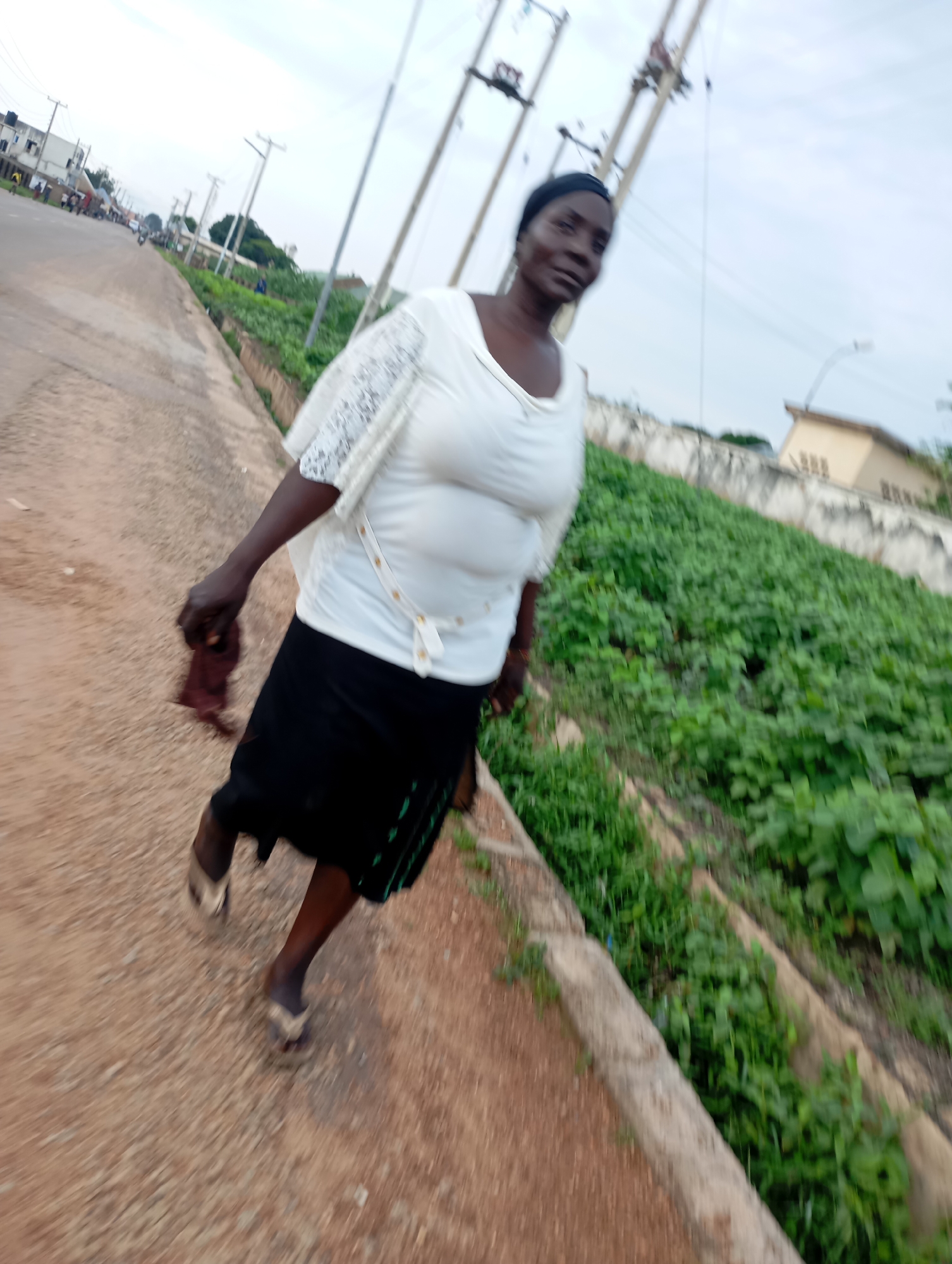 A full-shot, eye-level, medium-speed motion blur photograph captures a Black woman walking along a dirt path beside a road. The woman is wearing a white, short-sleeved shirt with lace accents on the sleeves and a black skirt. She has dark skin and is wearing a dark headscarf. She is holding a dark, folded cloth in her right hand. She is wearing flip-flops. To the woman's left is a paved road with a sidewalk. In the background, there are buildings, power poles, and a patch of green vegetation. The sky is overcast. The photograph was taken in Shendam, Nigeria.