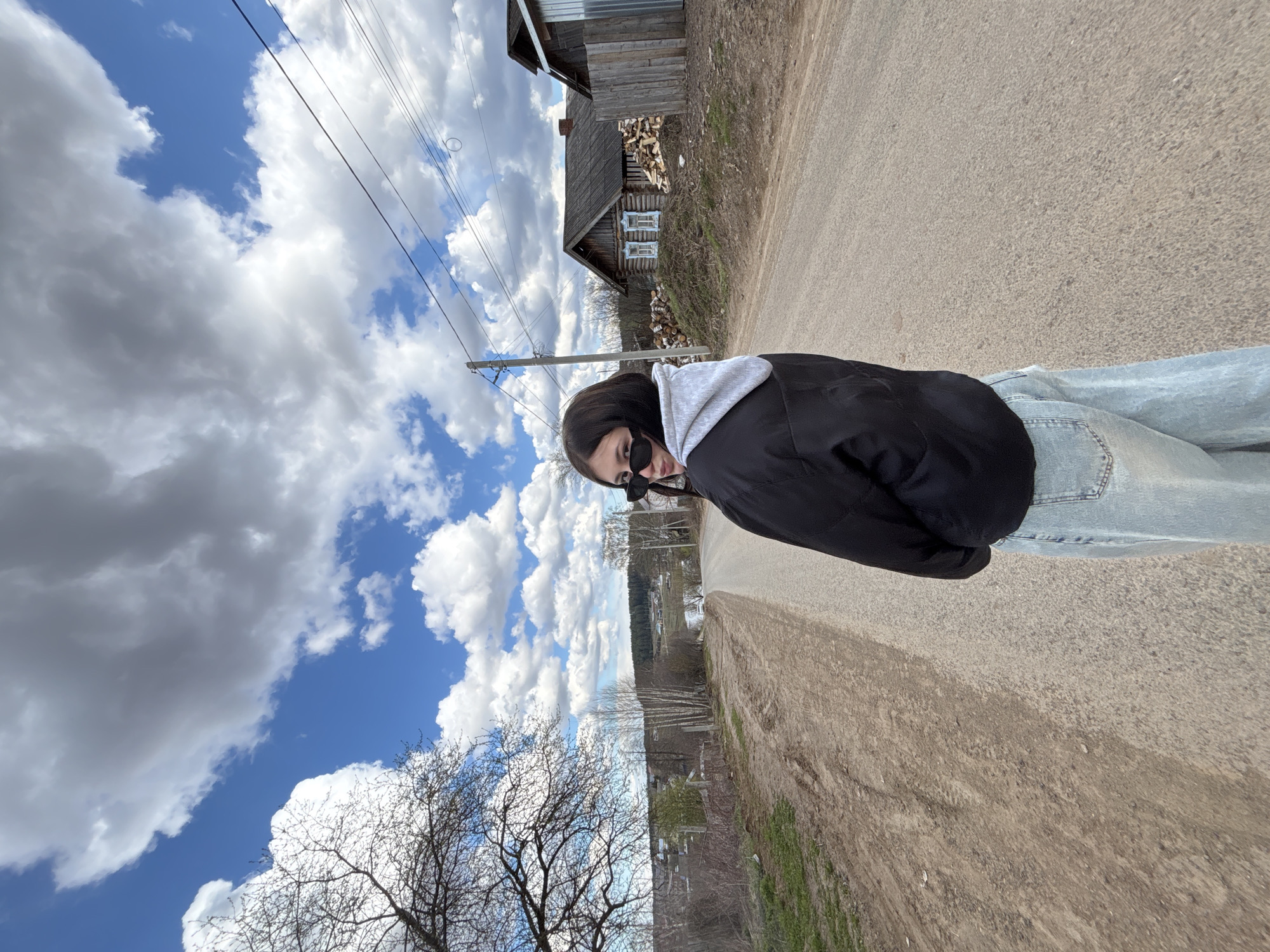The image displays a young woman standing on a rural road under a bright, partly cloudy sky. The original image is rotated 90 degrees counter-clockwise. The woman, the primary subject, has long dark hair, wears dark sunglasses, a black jacket with a light grey hood, and light blue jeans. She is looking directly towards the viewer with a casual pose.
The scene is a rustic outdoor setting, characteristic of a village. In the background, to the woman's right, is a weathered wooden house with blue window frames, and a stack of firewood is visible next to it. Utility poles and wires run across the upper mid-ground. The sky features prominent, fluffy white cumulus clouds against patches of clear blue. Bare-branched trees, possibly indicating early spring or late autumn, frame parts of the scene. The road surface appears to be a mix of worn asphalt and dirt/gravel. In the distance, a landscape with a body of water or valley and more trees can be seen. The bright daylight suggests it is daytime. No discernible text is visible in the image. The setting aligns with the provided location context of Фоки, Russia.
