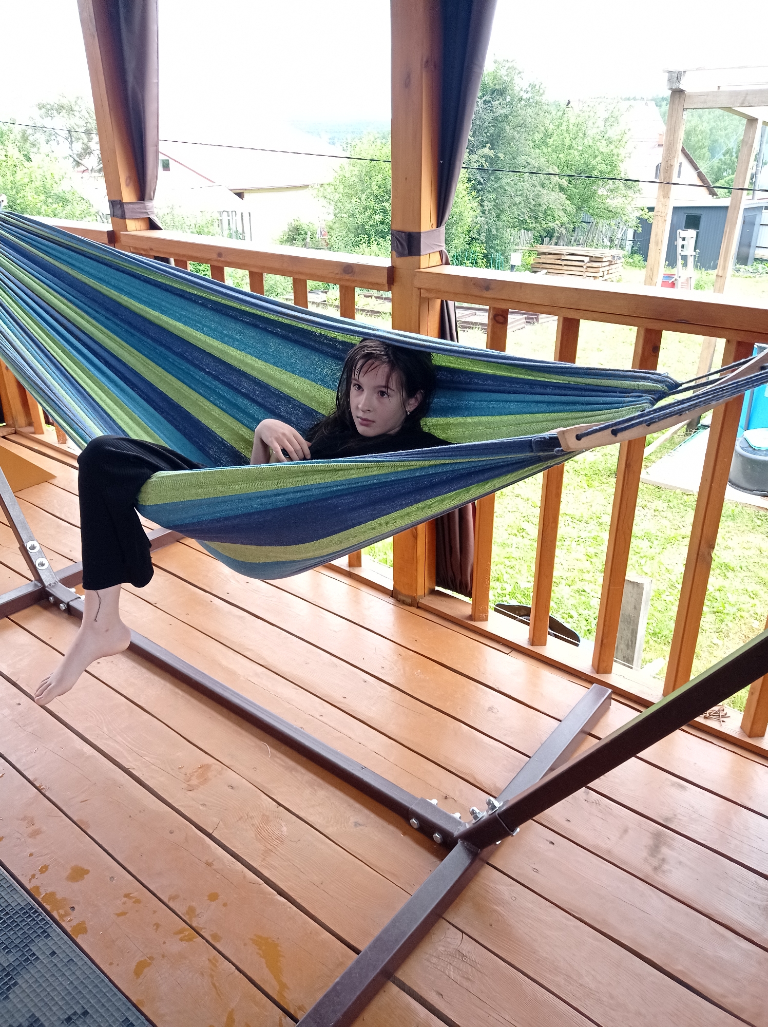 A young person with dark hair is lying in a striped hammock on a wooden deck. They are wearing black pants and a black top. Their left leg is extended out of the hammock, and their bare foot is visible, with a small tattoo on the ankle. The hammock has blue, green, and yellow stripes and is supported by a metal stand. The deck appears to be part of a porch or balcony, with wooden railings and support beams. In the background, there are trees, houses, and a cloudy sky. The setting suggests an outdoor recreational area, possibly a backyard or a campsite. The lighting indicates it is daytime, with overcast weather. The person's facial expression is neutral, suggesting relaxation or contemplation. No visible text is present in the image. The location context is Degtyarsk, Russia.