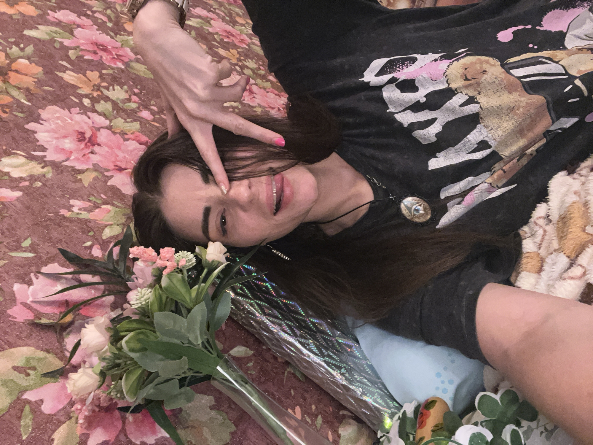 In Krasnoe-na-Volge, Russia, a woman with long dark hair and brown eyes is lying down on a bed with a floral patterned wallpaper in the background. She is wearing a black t-shirt with a graphic print. Her left arm is raised, with her fingers forming a peace sign over her eye. She is sticking her tongue out playfully. A bouquet of pink and white flowers in a clear vase is visible to her left, along with a pillow.