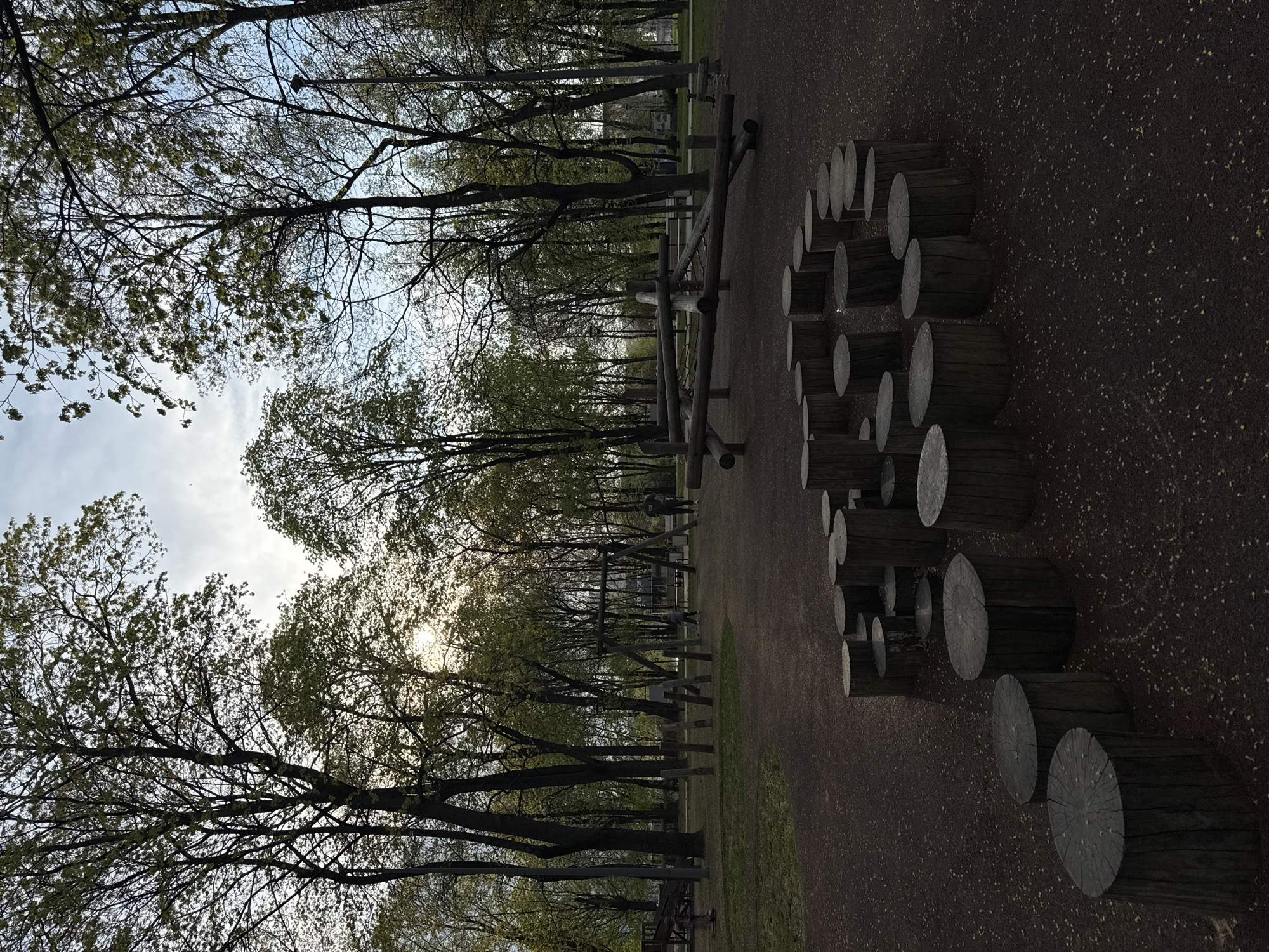 The image depicts an outdoor scene, likely a playground or park, possibly in Moscow, Russia. The foreground contains a series of wooden log rounds arranged in a curved pattern on a dark, textured ground surface. In the midground, there are wooden playground structures, possibly climbing frames or bridges, aligned in a row. Beyond the playground equipment are trees with sparse green foliage, and further in the background, the sky is visible with a mix of clear areas and some cloud cover. The scene is slightly overcast, giving the impression of late afternoon or early evening light.
