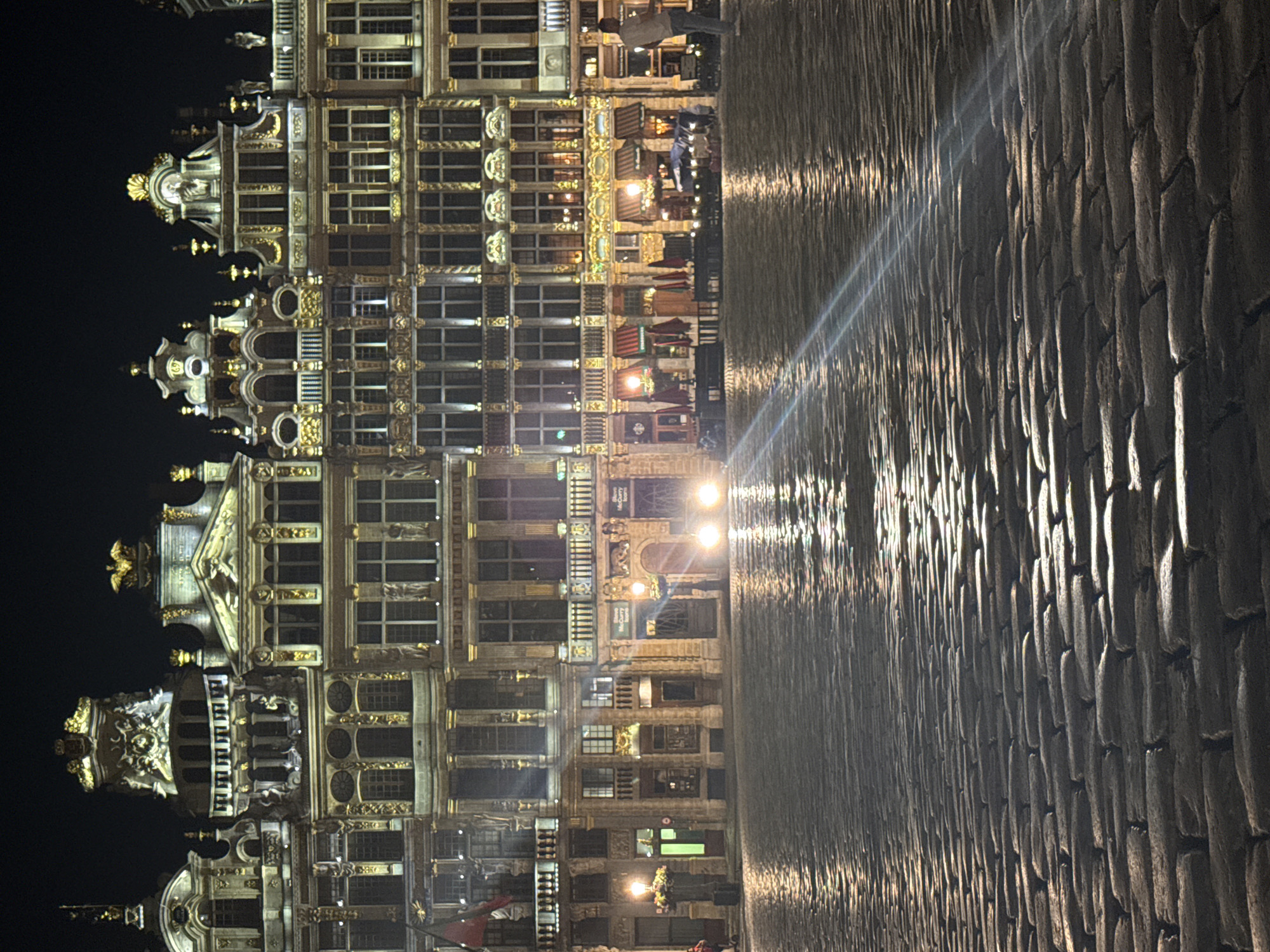 The image depicts the Grand Place in Brussels, Belgium, at night. The scene is illuminated by streetlights and the lights from the surrounding buildings, casting reflections on the wet cobblestone ground. The architecture of the buildings is ornate and historic, featuring elaborate facades with gold accents and numerous windows. Several businesses with awnings are visible on the ground floor of the buildings. In the foreground, the cobblestone pavement dominates the view, showing a texture of interlocking stones. In the background, headlights from two vehicles are visible in the distance, suggesting a street or road crossing the square. A few individuals are present, appearing as small figures in the vast open space. One person is standing near the edge of the buildings on the right side of the frame, and another person is partially visible on the left. The sky is dark, indicating nighttime.