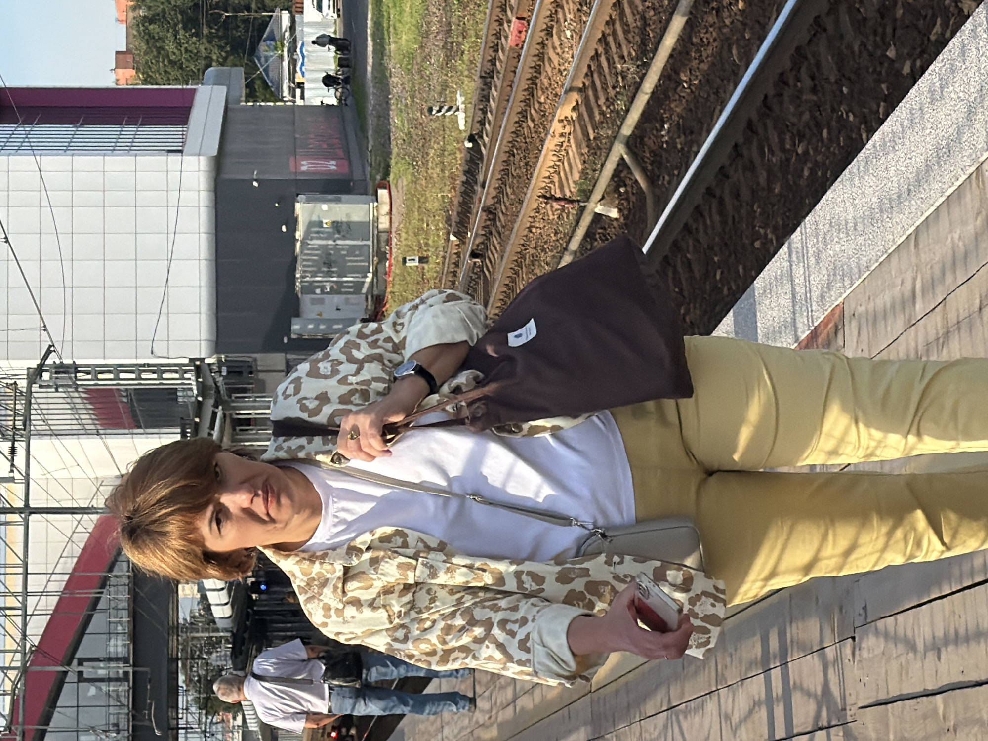 A woman with shoulder-length brown hair and a concerned expression walks along a wooden train platform. She wears a white t-shirt, a light-colored jacket with a tan and brown leopard print, and yellow pants. She carries a dark brown tote bag over her right shoulder and a small gray crossbody bag. Her left hand holds a smartphone. To her left, a man with gray hair, wearing a light blue short-sleeved shirt and jeans, walks away from the camera with a dark bag slung over his left shoulder. In the background, a modern building with white tiled walls and pink accents is visible, along with train tracks. The scene appears to be a train station in Shcherbinka, Russia, under a clear sky.
