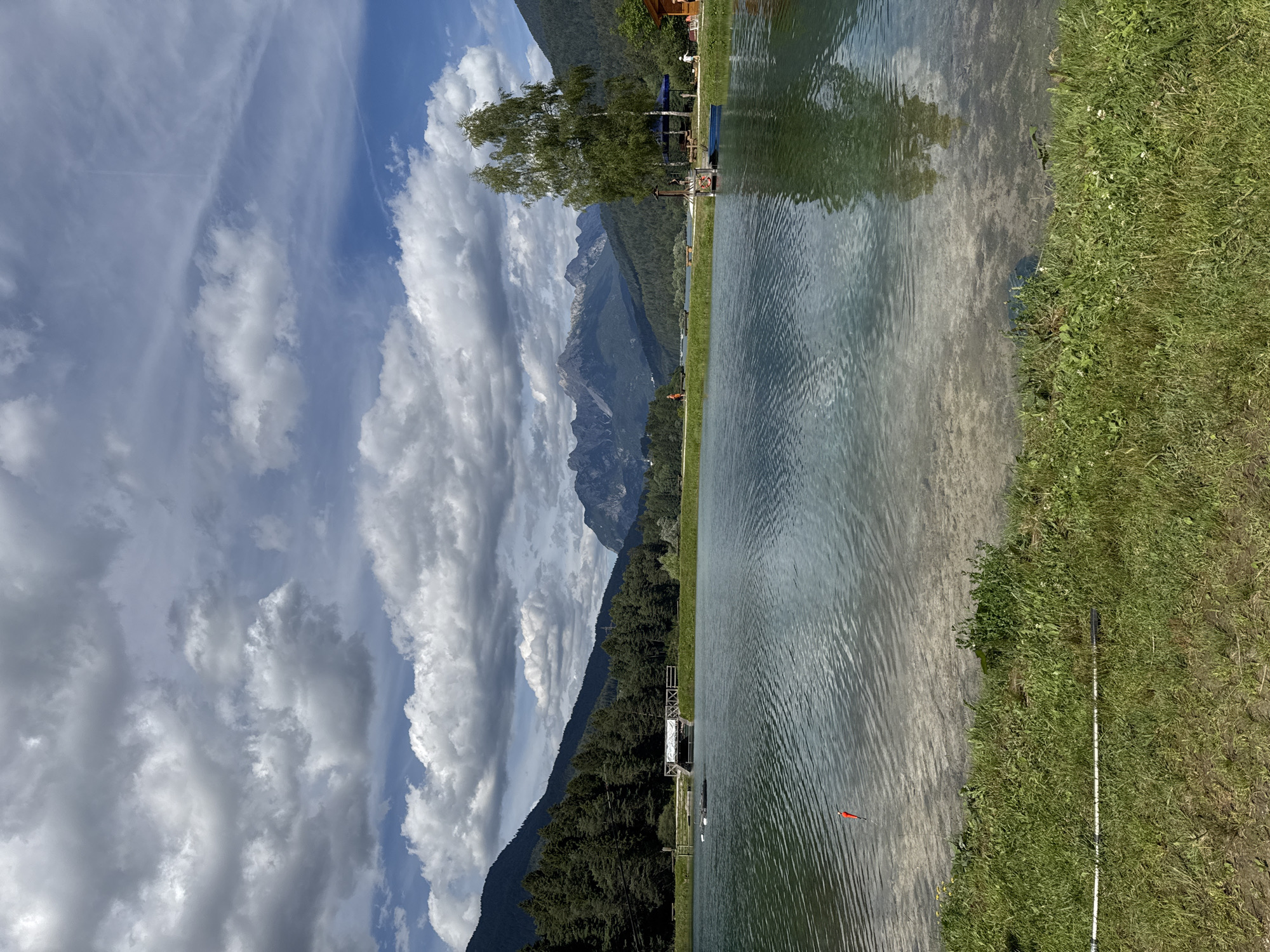 The image depicts a serene lakeside scene, likely in Calalzo di Cadore, Italy. The perspective is from the edge of a body of water, looking towards a tree-lined shore with imposing mountains in the background. The sky is a brilliant blue, adorned with dramatic, fluffy white clouds that cast shadows, suggesting it is daytime, possibly late morning or early afternoon.
The water's surface is rippled, reflecting the sky and the surrounding greenery. A sandy bottom is visible in the shallow areas near the shore. On the right side of the frame, a lush green grassy bank slopes gently towards the water. A long, white object, possibly a fishing rod or a surveying pole, lies horizontally on the grass, near the water's edge. Further out in the water, a small red marker float is visible.
The left side of the image features a dense forest covering the lower slopes of the mountains. The peaks of the mountains are jagged and rocky, reaching towards the sky. A tall tree with green foliage stands prominently on the shore, casting a shadow. In the distance, a small structure or building can be seen on the shoreline, along with what appears to be a pier or platform extending into the water. A few indistinct figures are visible near the shore and on the platform, engaged in leisure activities, though their specific actions are not discernible. The overall impression is one of a peaceful, idyllic natural setting, bathed in sunlight and offering opportunities for recreation. There is no visible text in the image.