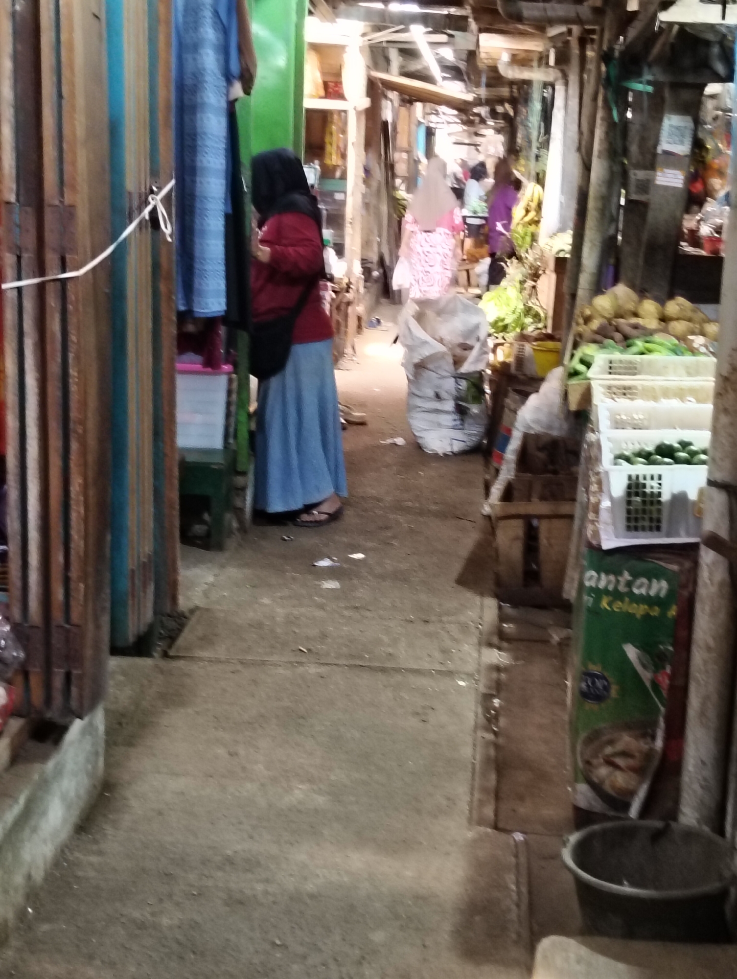 The image depicts a market scene in Subah, Indonesia. In the foreground, a woman wearing a red hijab and a long blue skirt stands near a stall. She is partially obscured by vertical wooden planks. To her right, a narrow walkway leads into the market. Further into the market, several other individuals are visible in the background, some wearing hijabs and colorful clothing. Stalls are laden with produce, including what appear to be coconuts and green vegetables displayed in white plastic crates. Large white sacks are also present, suggesting goods being sold or stored. The environment appears to be an open-air market with wooden structures and a concrete floor. The overall lighting suggests it may be late afternoon or early evening.