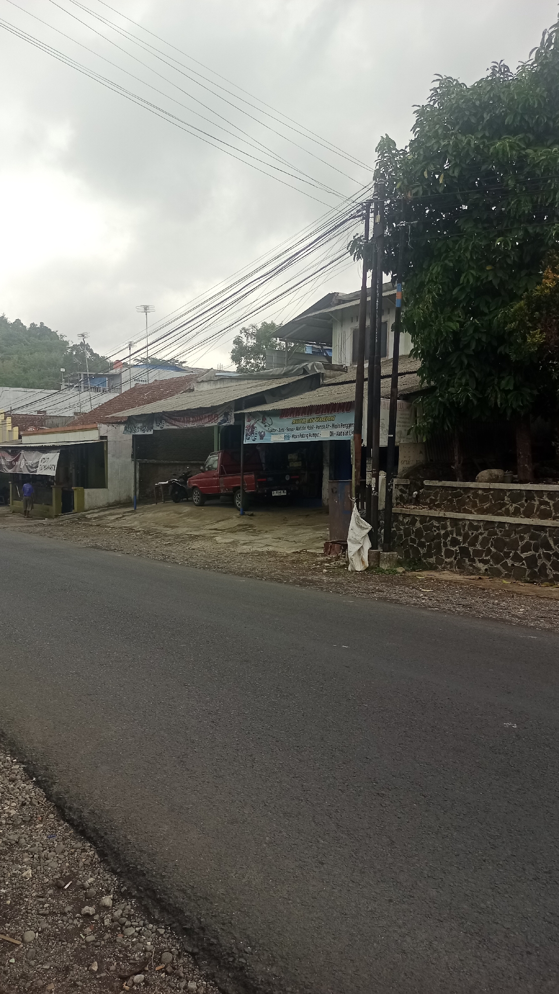 The image depicts a street scene in Karyamandala, Indonesia, under an overcast sky. In the foreground, a paved road runs diagonally from the bottom left towards the center of the frame. The road is bordered on the left by a gravelly shoulder.
On the right side of the road, a building with a corrugated metal roof is visible. It appears to be a commercial establishment, possibly a workshop or repair shop. A red pickup truck is parked under the shelter of the building, and a motorcycle is also present. There is a banner hanging from the building with Indonesian text and graphics. Two utility poles with numerous wires are situated in front of the building. A white plastic bag is tied to one of the utility poles. Behind the building, a stone wall is constructed.
Further back, to the left of the commercial building, are other structures with corrugated metal roofs and some houses. A person in a blue shirt is standing outside one of these structures. In the background, a hill with vegetation is visible. The overall impression is of a semi-urban or semi-rural street in a tropical climate.