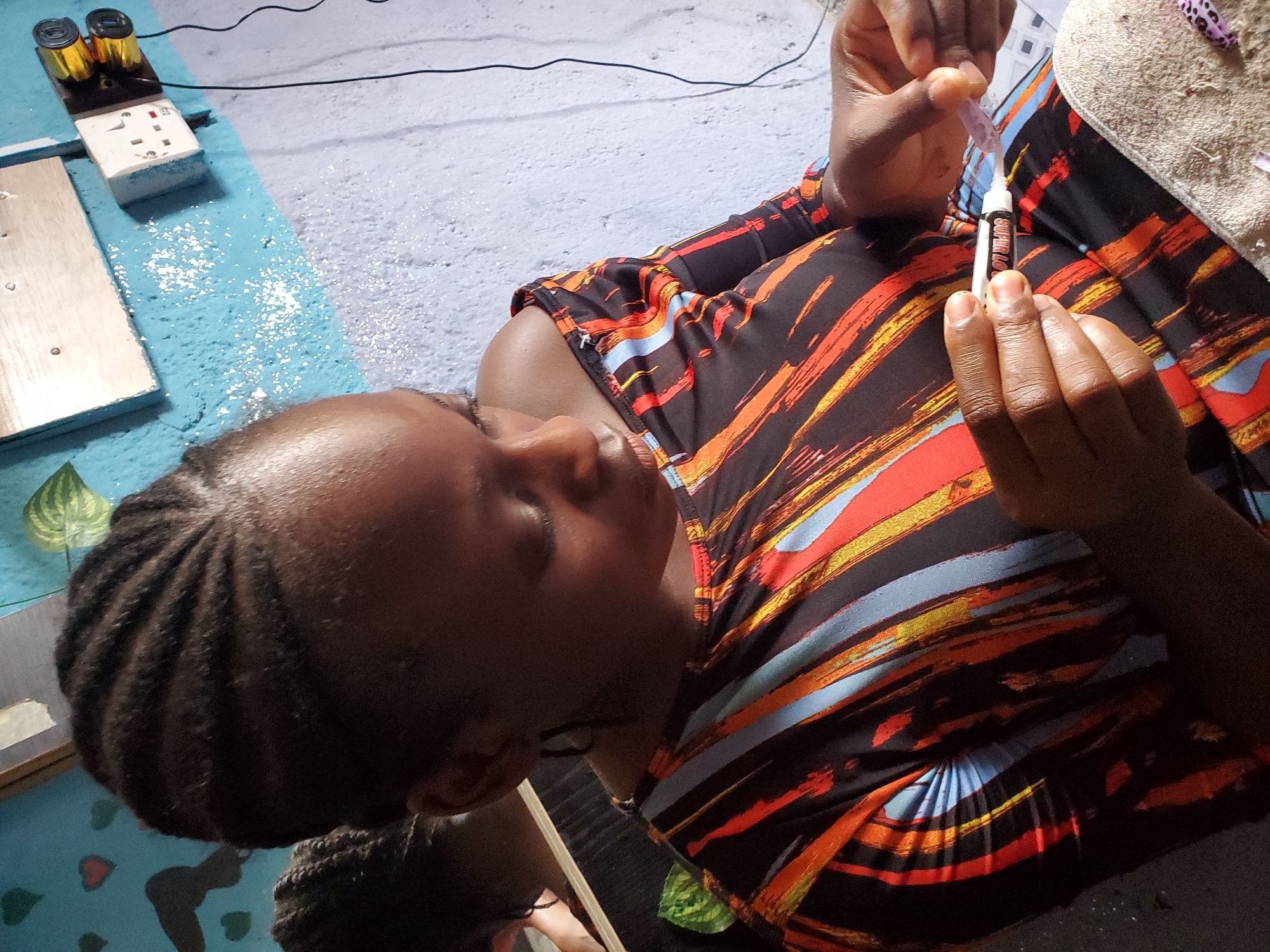 The media file shows a close-up, overhead view of a young Black person, likely a child or young teenager, engaged in an activity. The person is wearing a colorful shirt with a black base and vibrant, abstract streaks of orange, yellow, and blue. Their dark hair is styled in neat braids.
The person is holding a small tube of what appears to be glue or a similar adhesive in their right hand, with the nozzle directed towards their left hand. The left hand is holding a small, translucent purple object, possibly an artificial nail or a craft item, to which the adhesive is being applied. The person's head is tilted down, focused intently on the task. Their expression is one of concentration.
The setting appears to be indoors, on a textured, light-colored surface. To the left of the person, a blue surface with white speckles is visible, and on it sits a white electrical socket with two gold-colored cylindrical objects, possibly batteries, attached to a black base. A thin black wire extends from this assembly. A wooden plank is also visible on the blue surface. A beige towel or cloth is draped to the right of the person.
The lighting suggests it is daytime, with natural light casting shadows. The overall mood is one of quiet concentration and creative engagement. No specific time of day, weather, or explicit emotions beyond focus are evident. There is some visible text on the white glue tube, but it is largely illegible in the image. The location context is Jos, Nigeria.