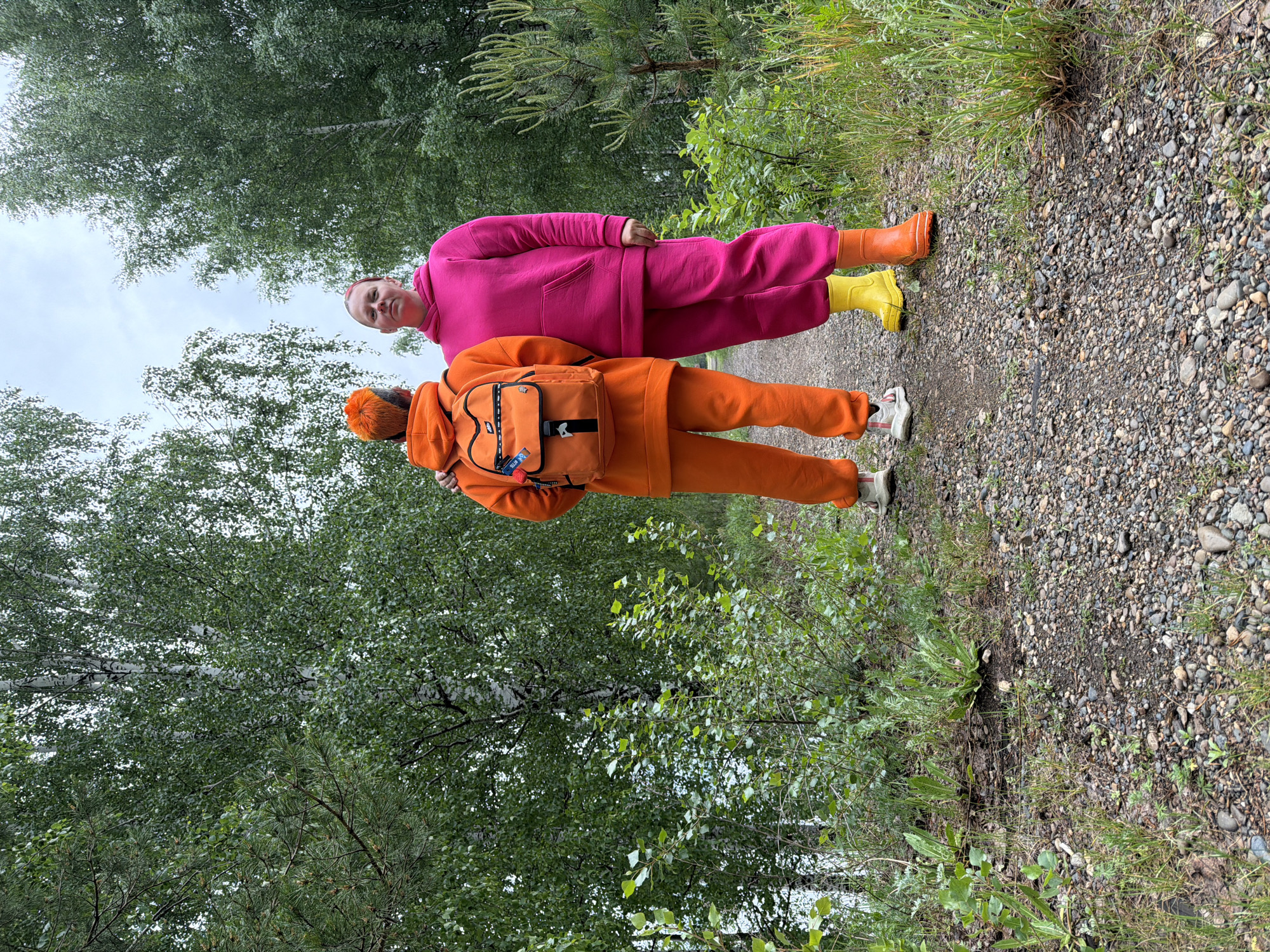 Two individuals stand on a gravel path in a wooded area. One person, wearing a hot pink sweatsuit and yellow and orange rain boots, faces the camera with a neutral expression. The other person, dressed in an orange sweatsuit and white sneakers, has an orange backpack. The path is surrounded by trees and grass, suggesting a park or forest setting. The location is Chaikovsky, Russia.