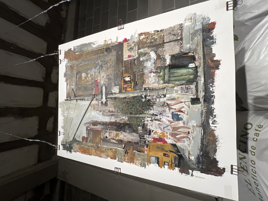 A large, unframed, vertically oriented artwork depicting a textured, impressionistic urban street scene is suspended in an industrial or storage environment. The artwork portrays a city street corner with several multi-story buildings, a partial view of a yellow van with a partially visible license plate ("H377") on the left, and a group of approximately five to seven pedestrians walking across a crosswalk or sidewalk in the foreground. A street pole featuring a pedestrian traffic signal displaying a white "walk" symbol and an illegible street sign is central on the right. Below the signal, two dark green waste bins are positioned near a chain-link fence. Overhead, utility lines crisscross the upper portion of the painted cityscape.
The artwork is secured by four metal binder clips attached to its top and bottom edges, from which thin wires extend upwards, suspending it from an unfinished concrete slab ceiling with exposed joists. To the left of the artwork, a raw concrete column is visible, while a gray cinder block wall is present to the right. Directly beneath the artwork, several large white bags are stacked, with text "EL ENCINO" and "beneficio de café" clearly visible on one of them. This suggests the setting is a coffee processing or storage facility located in the city of León, Mexico.