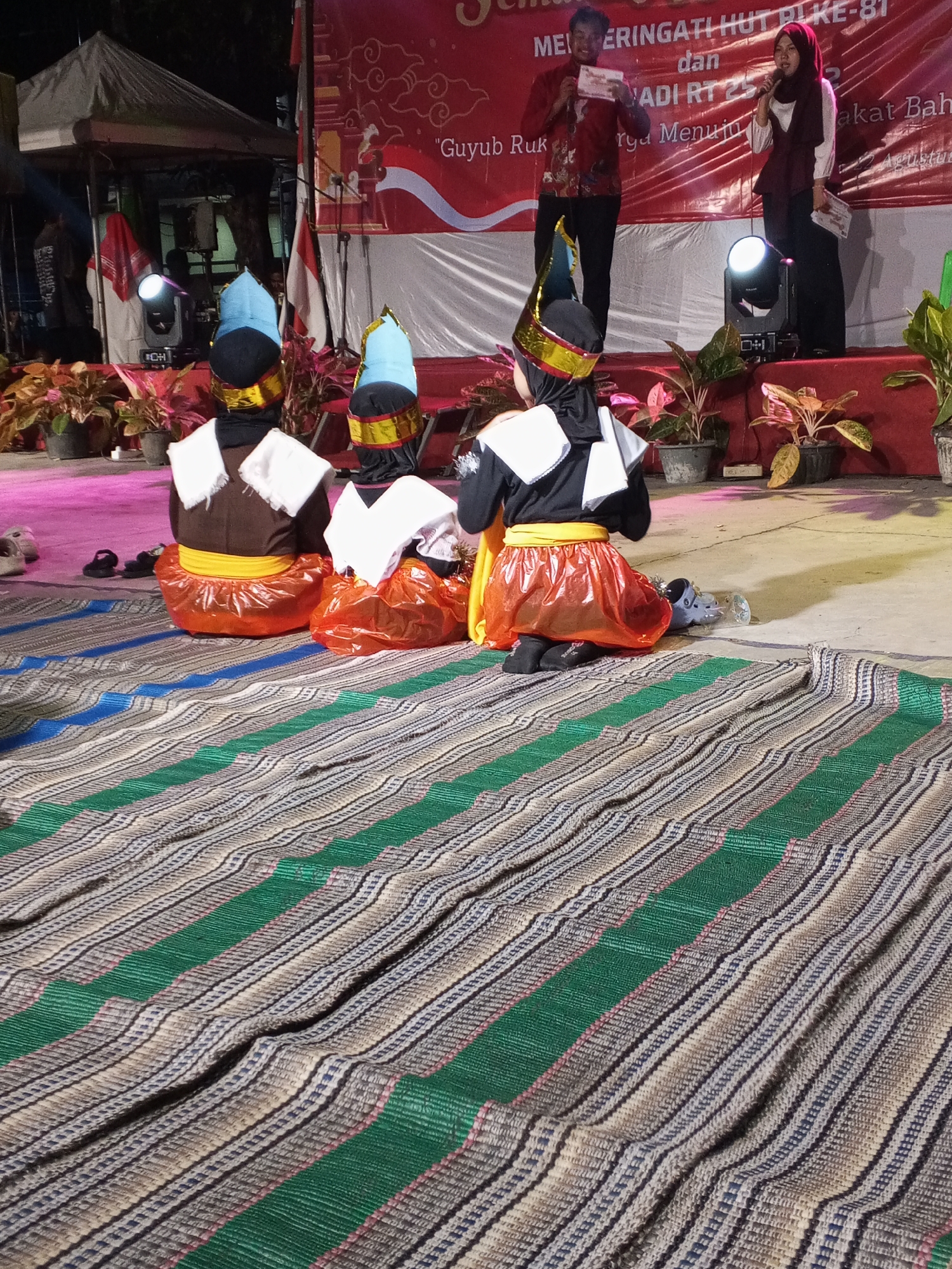 The image depicts an outdoor stage event in Ciruas, Indonesia, likely during an evening celebration, given the artificial lighting. In the foreground, three individuals, likely children, are seated on a woven mat, facing away from the camera. They are dressed in traditional performance attire, including brightly colored conical headdresses, brown tops, yellow sashes, and iridescent orange skirts that appear to be made of plastic material. Their backs are to the audience, suggesting they are either performing or waiting for their turn.
Behind them, on a raised stage, two adults are visible. A man in a red patterned shirt and black pants stands to the left, holding a white paper. To his right, a woman wearing a hijab and a maroon top with a white undershirt speaks into a microphone. A banner with Indonesian text, partially obscured by the performers, hangs on the stage backdrop. The stage is decorated with potted plants and illuminated by stage lights. The overall setting suggests a community event or festival.