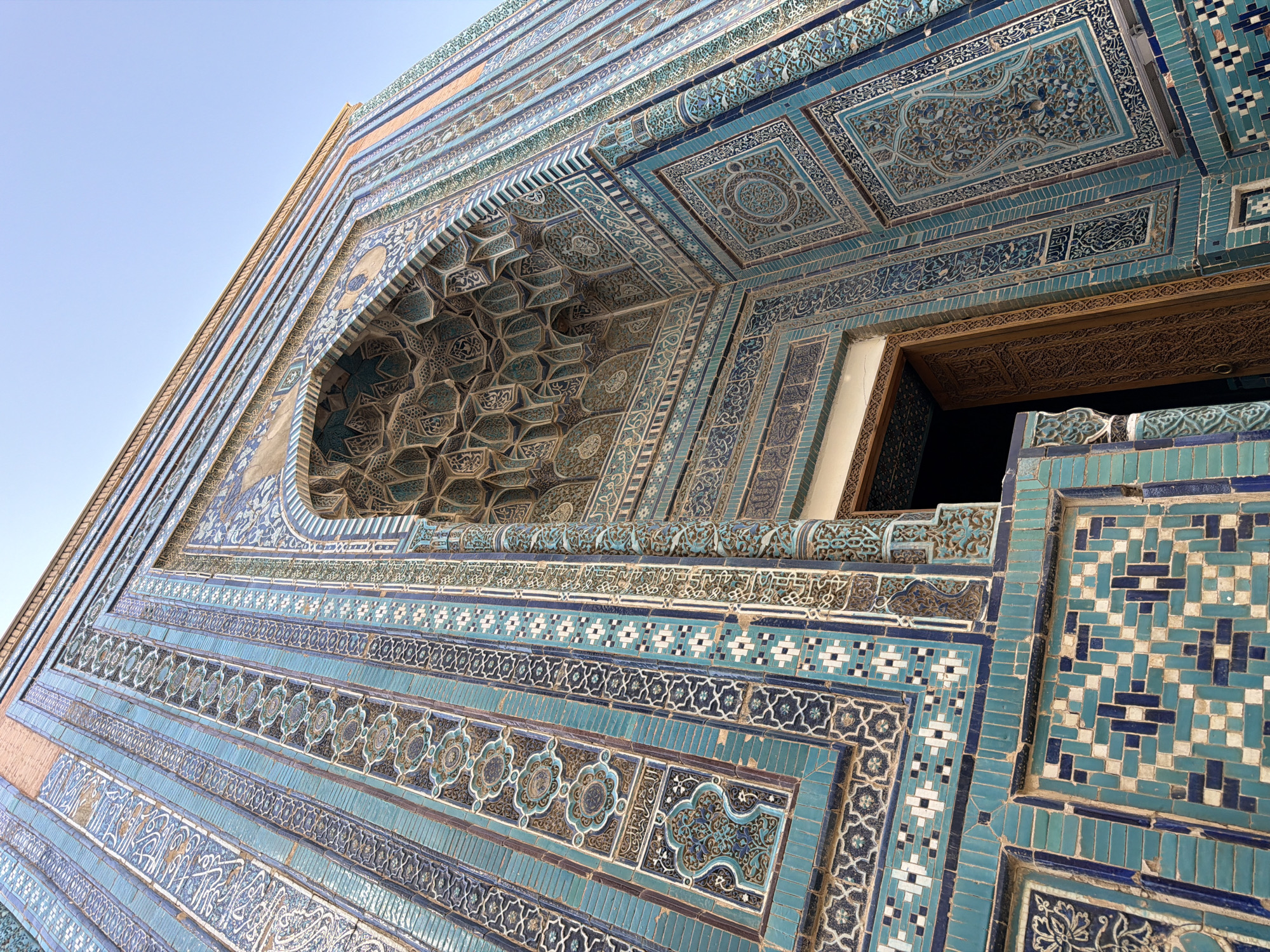 The image shows a low-angle, upward view of the exterior of a building adorned with intricate blue and turquoise tilework. The building features a large, deeply recessed archway, the soffit of which is covered in a complex geometric pattern of muqarnas. Flanking the archway and extending along the facade are vertical bands of decorative tiles featuring Arabic calligraphy, floral motifs, and geometric designs in shades of blue, turquoise, white, and brown. The entrance beneath the archway appears to be a wooden door with carved detailing. The sky above is clear and pale blue. This architectural style is characteristic of the Shah-i-Zinda necropolis in Samarkand, Uzbekistan, a UNESCO World Heritage site known for its stunning mausoleums and tilework. There are no people or animals visible in the frame.