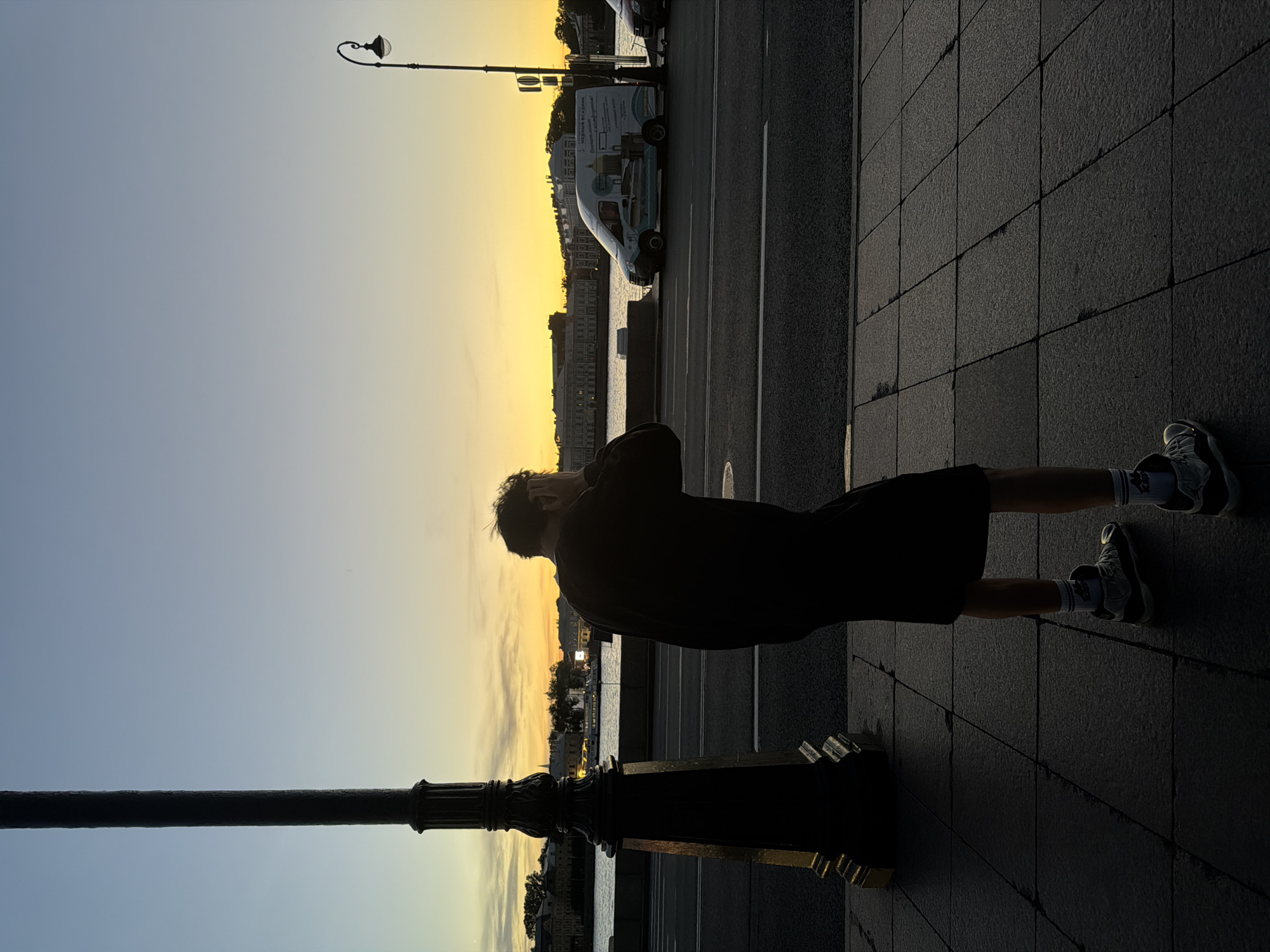 The image depicts a person standing on a sidewalk in Saint Petersburg, Russia, during sunset. The person is silhouetted against the warm glow of the setting sun, with their hands covering their ears. They are wearing a dark shirt, shorts, and sneakers with white socks. To the left, a tall, ornate lamppost stands prominently, also in silhouette. In the background, across a body of water, buildings line the embankment, and a white van is parked on the road. The sky transitions from a pale blue at the top to a vibrant yellow and orange near the horizon, indicating the time of day. The sidewalk is paved with large, dark tiles. The scene suggests a moment of quiet contemplation or distress for the individual.