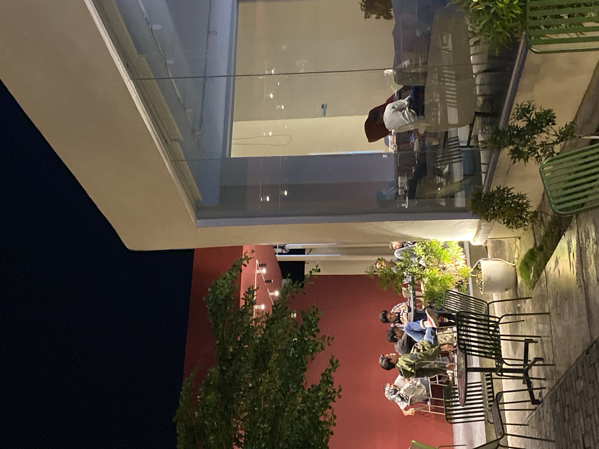 This media file captures an evening scene at an outdoor seating area, likely part of a cafe or restaurant in Lhokseumawe, Indonesia. The image is oriented vertically but depicts a horizontal ground level.
**Subjects:** The primary subjects are a group of young people seated at black slatted metal tables and chairs on a paved patio. They appear to be casually dressed, engaged in conversation, using mobile phones, or simply relaxing. More indistinct figures are visible through the large glass windows of an upper floor, seemingly inside the building. Several green plants, both in pots and planted directly, are interspersed throughout the seating area, adding a touch of nature. Empty black and green slatted chairs are also visible.
**Activity or event taking place:** The image depicts a social gathering at a relaxed outdoor establishment during the night. People are spending leisure time, interacting, and enjoying the evening atmosphere.
**Notable details:** The time of day is clearly night, indicated by the dark sky and the reliance on artificial lighting. The patio area is illuminated by lights mounted on a prominent red wall in the background, and by ambient light emanating from the building's interior, visible through its large glass windows. The building itself features modern architecture with a cream-colored facade on the upper floor and extensive glass panels that reflect interior lights and subtle details of the outdoor environment. The furniture includes contemporary black metal slatted chairs and tables, along with some green slatted chairs. The ground is paved, suggesting an urban or commercial setting. The overall mood appears calm and social. No visible text is present in the image.