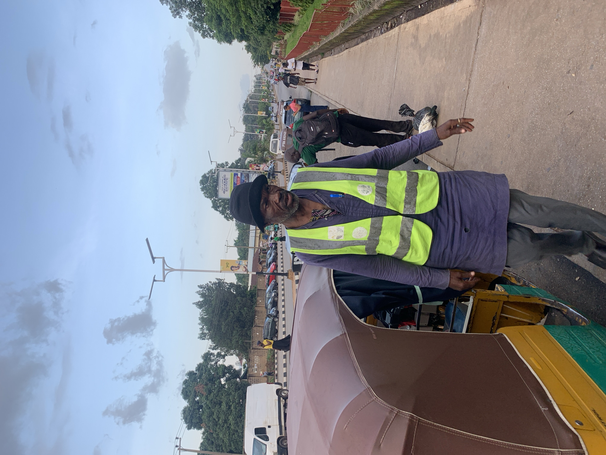 A man wearing a black hat and a high-visibility vest walks on a sidewalk next to a street in Barakin Lerawi, Nigeria. He is in the foreground of the image, with a brown and yellow auto-rickshaw to his left. The sidewalk is made of concrete and runs alongside a street with traffic, including cars and a white van. In the background, trees line the street, and a few people are visible walking along the sidewalk. The sky is partly cloudy. The man in the foreground appears to be looking towards the right side of the frame. There is a black trash bag on the sidewalk near the man.