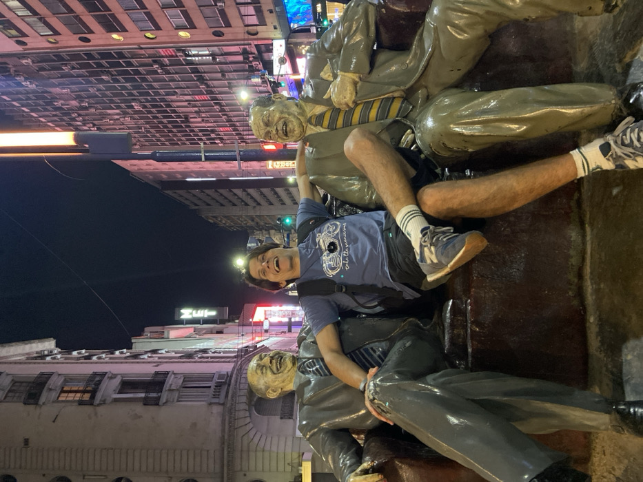 A man sits on a bronze statue of businessmen, arms raised. The statue, featuring caricatured faces and outdated suits, has a shiny, wet-looking surface. The man wears a blue t-shirt, dark shorts, athletic socks, and New Balance sneakers. A city street with tall buildings and neon signs provides the backdrop, illuminated under a night sky. The image appears rotated counter-clockwise.