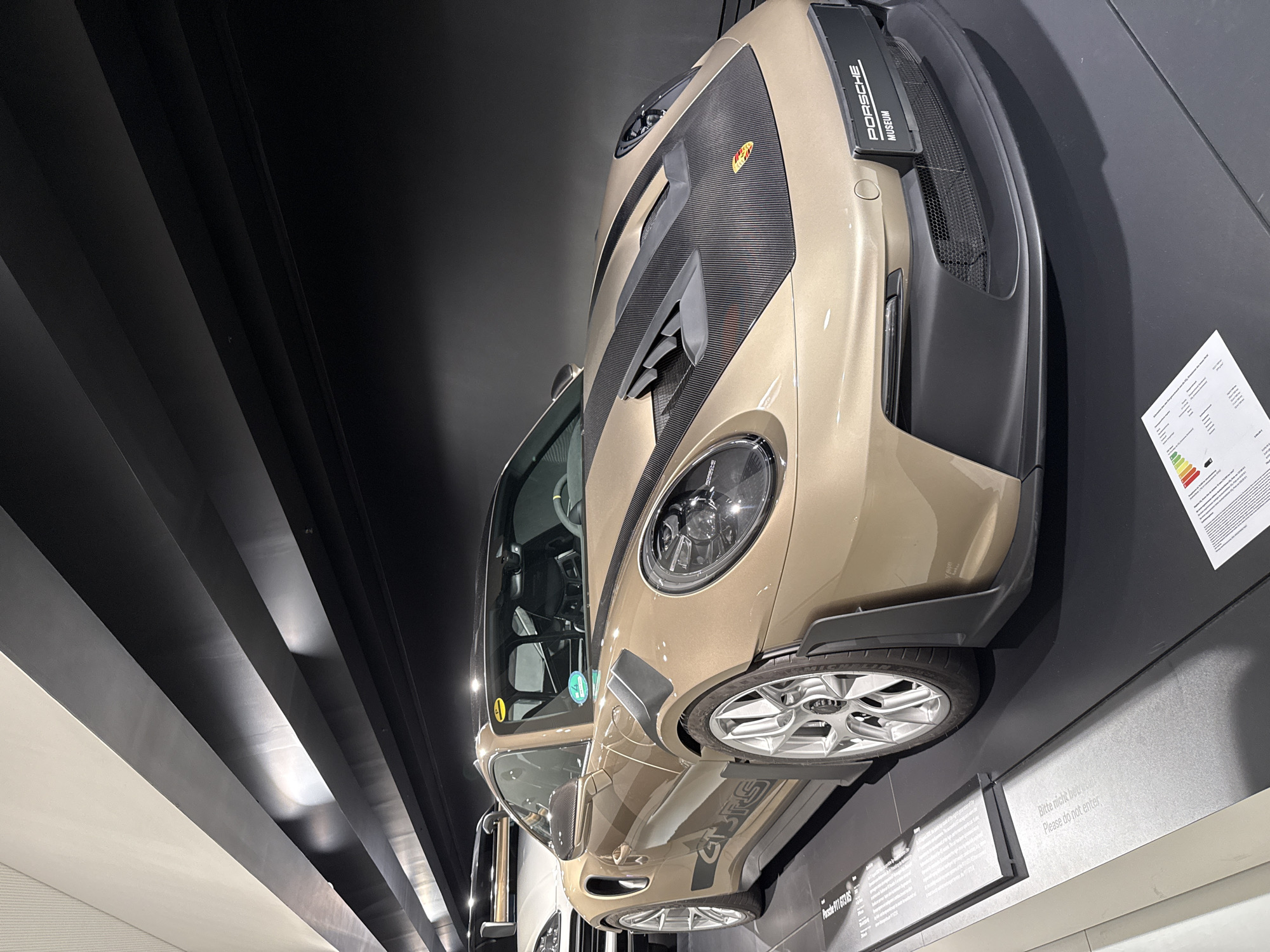 The image shows a gold-colored Porsche 911 GT3 RS on display at the Porsche Museum in Stuttgart, Germany. The car is positioned at an angle, with its front facing towards the right of the frame. It features a carbon fiber hood with prominent vents, large alloy wheels, and racing decals on its side. The car is situated on a black display platform. In the background, the museum's architecture is visible, characterized by dark, angled ceiling elements and spotlights. A white Porsche is partially visible behind the gold car. A sign reading "PORSCHE MUSEUM" is mounted on the front of the gold car. A printed document with energy efficiency ratings is placed on the floor in front of the car. The lighting in the museum is ambient and focused on the displayed vehicles.