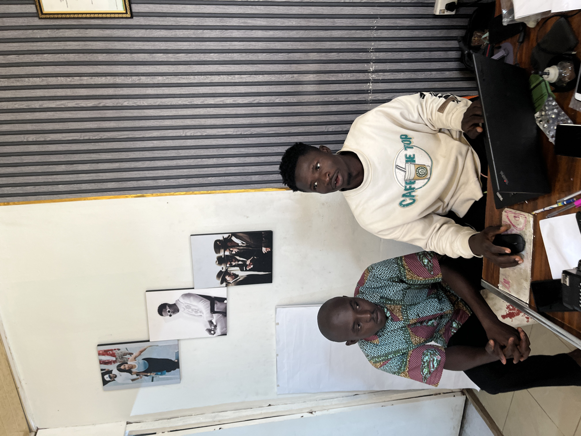 The image depicts two young men sitting at a table in what appears to be an office or creative space. The man on the left wears a colorful, patterned shirt and black trousers. He is looking down with his hands clasped in front of him. The man on the right is wearing a cream-colored sweatshirt with a "CAFFEINE TOP" graphic. He is looking to his right and has his hand on a laptop.
The environment features a wall with vertical wooden slats behind the man on the right, and a plain, light-colored wall on the left. Several framed pictures are hung on the left wall, including images of people in fashion or lifestyle poses. There is also a whiteboard visible. On the table in front of the men are a laptop, a computer mouse, pens, and other office supplies. The scene has natural lighting. The question about the specific location in Jos, Nigeria is not answerable from the visual information alone.