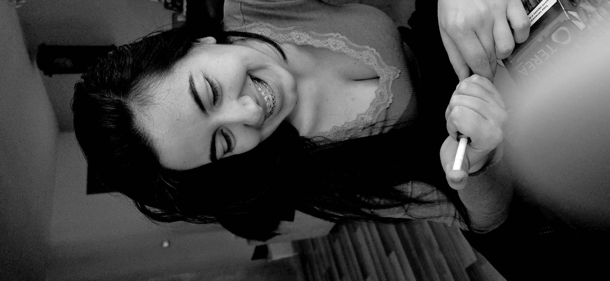 A black and white photo shows a woman with dark hair and braces, lying down with her eyes closed, smiling. She wears a top with lace trim. In the foreground, a hand holds a lit cigarette near a "OTEREA" package. The exact location in Varna, Bulgaria is not discernible from the image.