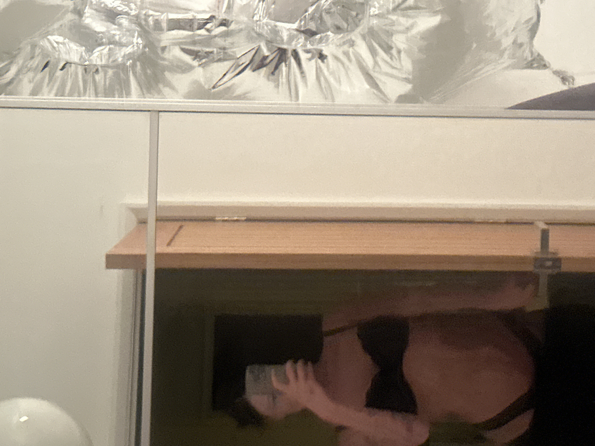 The image captures an indoor scene in Tunbridge Wells, United Kingdom, depicting a mirror selfie. A person with dark, short hair is reflected in a mirror, holding a smartphone that obscures their face. The individual is wearing a black bra and black lower undergarments. To the reflected person's right, a light-colored wooden doorframe is visible, indicating an open or partially open doorway. The reflection also shows a segment of a light-colored wall. Above, a spherical white light fixture is partially visible on the ceiling. To the far right of the image, an irregular, textured, silvery reflective surface, possibly crinkled metallic foil or material, is present, showing various reflections and creases. The overall lighting is subdued.