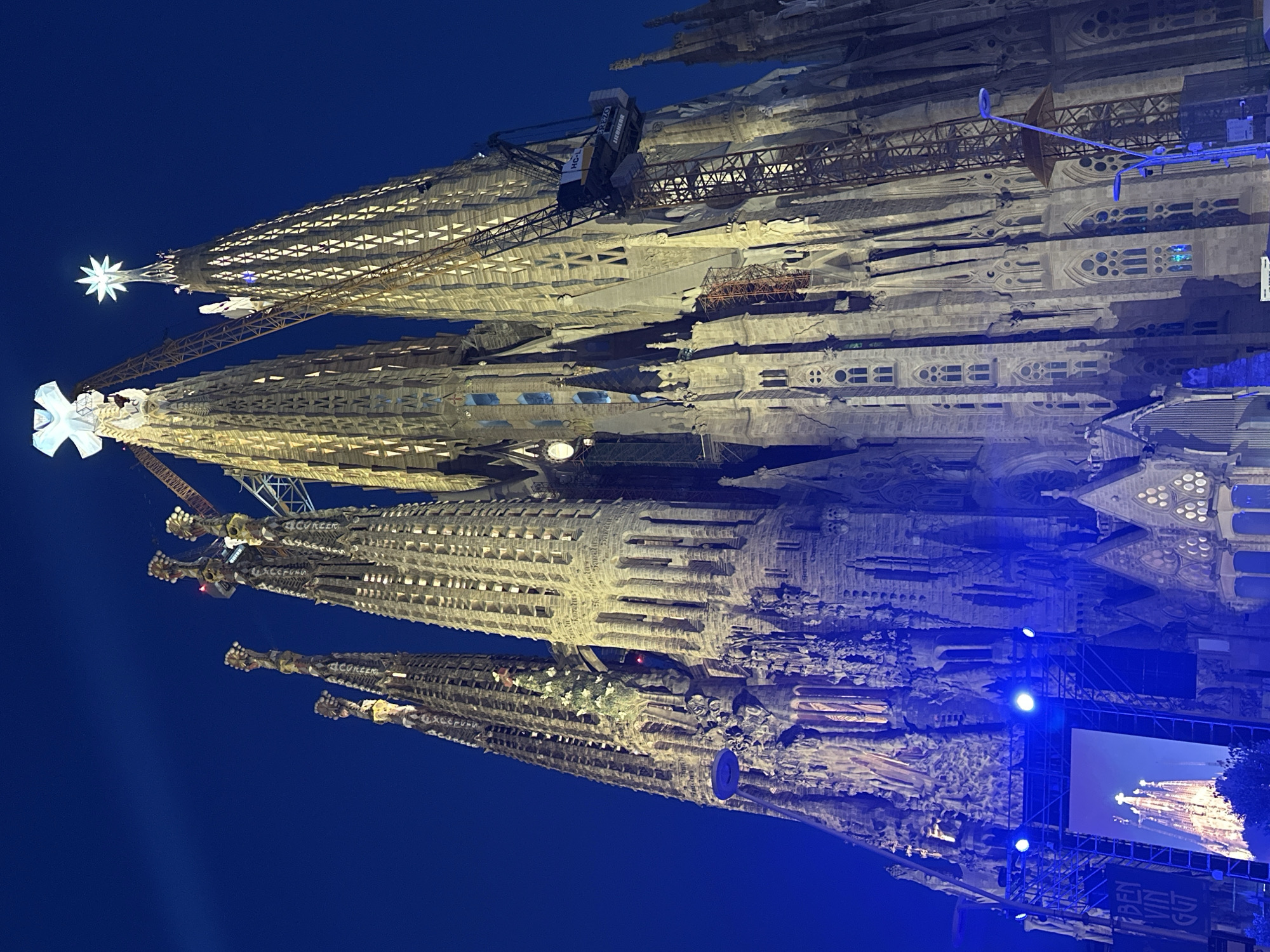 The image shows the Sagrada Familia basilica in Barcelona, Spain, at night. The basilica's iconic towers are illuminated, with a large star visible atop one of the spires and a cross atop another. Cranes are present, indicating ongoing construction. In the foreground, a large screen displays an image of the basilica, surrounded by stage lighting and scaffolding. The sky is a deep blue, suggesting dusk or nighttime. The overall scene captures the architectural grandeur of the Sagrada Familia while also highlighting its continuous development.