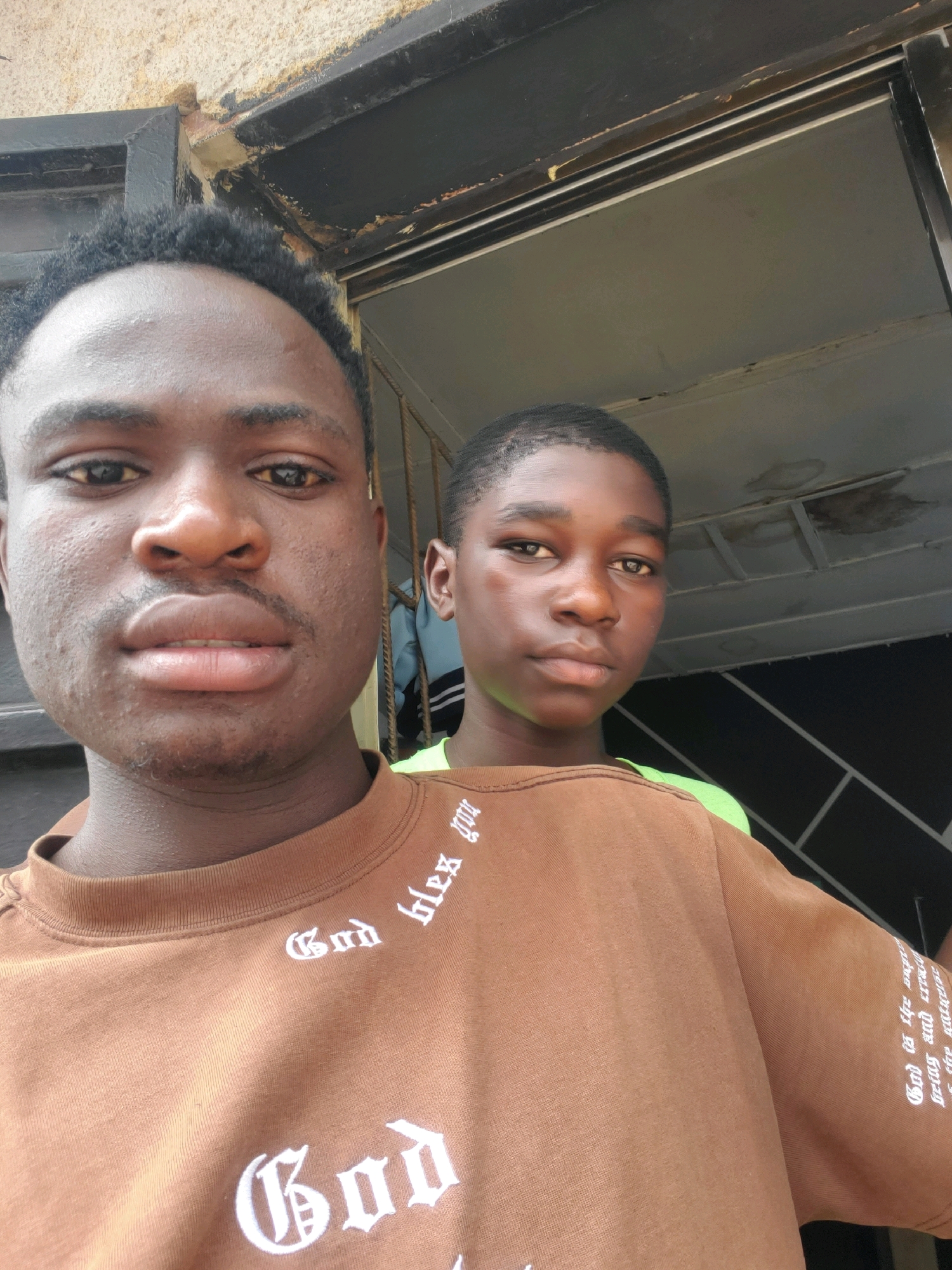 Two young males are positioned in the foreground of the image, facing the camera. The male on the left, closer to the camera, is shown from the chest up, with his head slightly tilted. He has dark skin, short black hair, and is wearing a brown t-shirt with white gothic-style lettering on the chest and sleeves. Visible text on the shirt includes "God" and "bless you". The male on the right, further from the camera, is also visible from the chest up. He has dark skin and short black hair, and is wearing a brown t-shirt with a bright green undershirt or strap visible around the collar area. The background suggests an outdoor setting, possibly a doorway or entrance. The wall above and to the left of the subjects appears to be textured and a light beige color, with a dark, possibly metal, frame above it. To the right of the subjects, there is a glimpse of what could be a building interior or a partition with a geometric pattern. The overall lighting suggests it is daytime. The image was taken in Jos, Nigeria, although specific landmarks or contextual clues within the image do not pinpoint the exact location within the city.