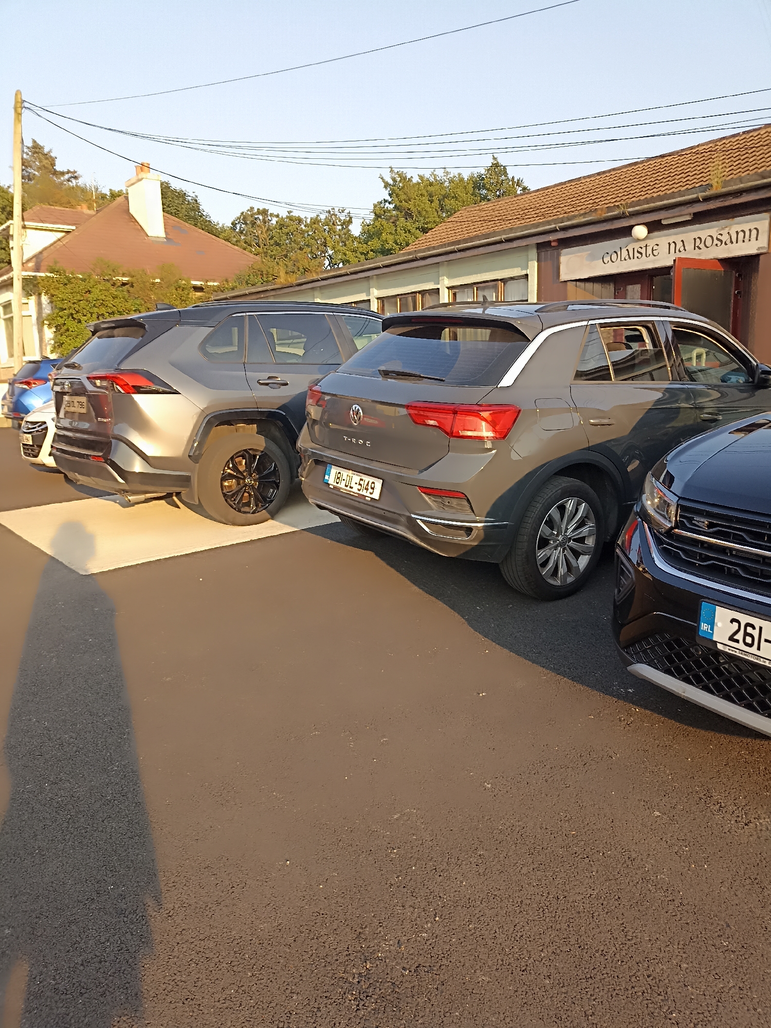 In Annagry, Ireland, on a paved surface near a building with a sign that reads "colaiste na rosann," several parked cars are visible. The cars include a grey Toyota RAV4, a grey Volkswagen T-Roc, and a black Volkswagen, all facing away from the camera. The RAV4 has a license plate that reads "191-DL-796," and the T-Roc has a license plate reading "19-DL-5149." The black Volkswagen's license plate is partially visible, showing "261-". A shadow of a person extends across the pavement in the foreground, cast by the setting sun. The building appears to be a school or community center.