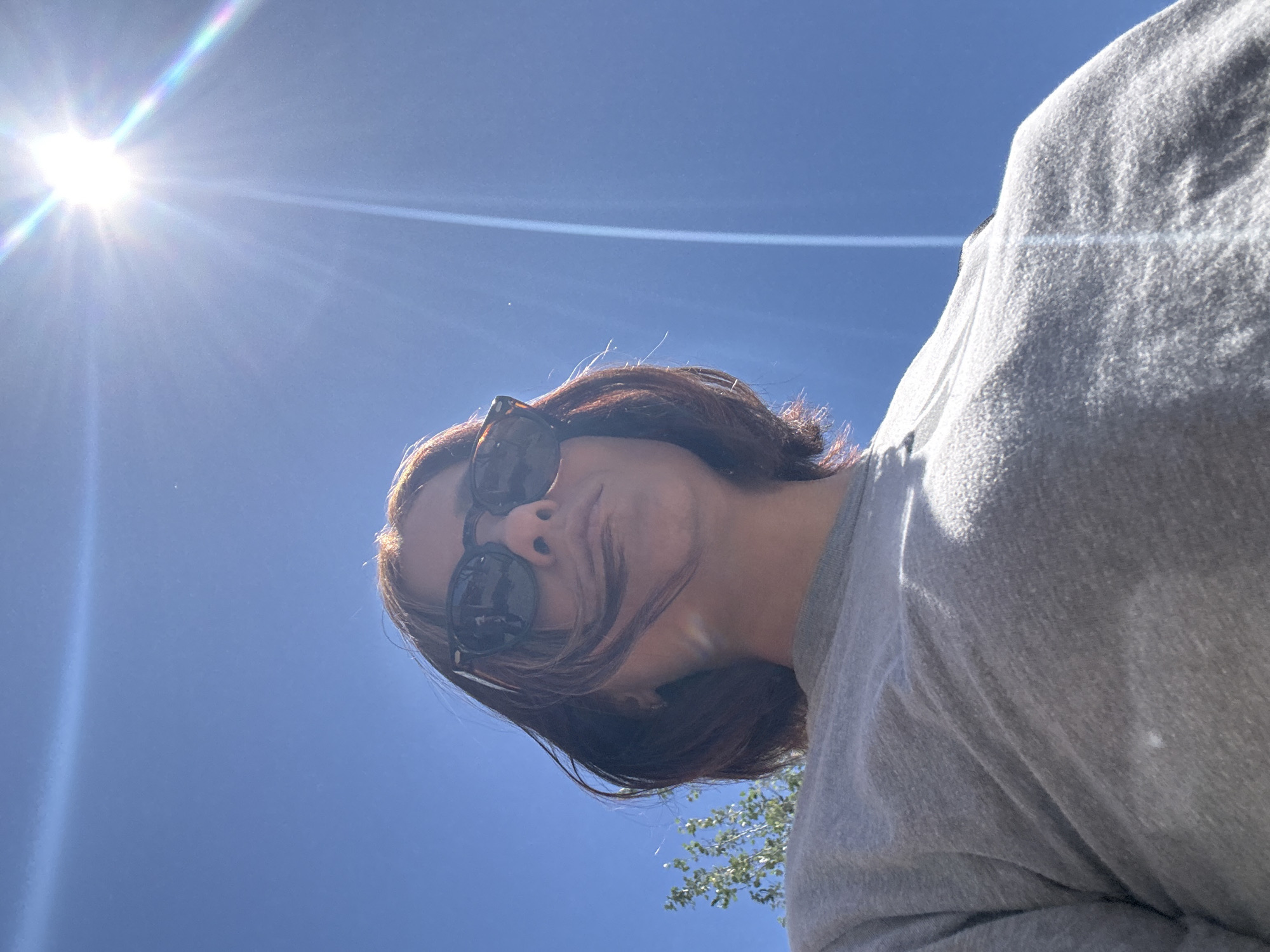 A low-angle, outdoor shot depicts a person wearing sunglasses and a grey, long-sleeved shirt, set against a clear blue sky with a bright sun visible in the upper left corner. The person's brown hair is partially obscuring their face. Some foliage is visible at the bottom of the frame. The location cannot be determined to be Barcelona from this image alone.