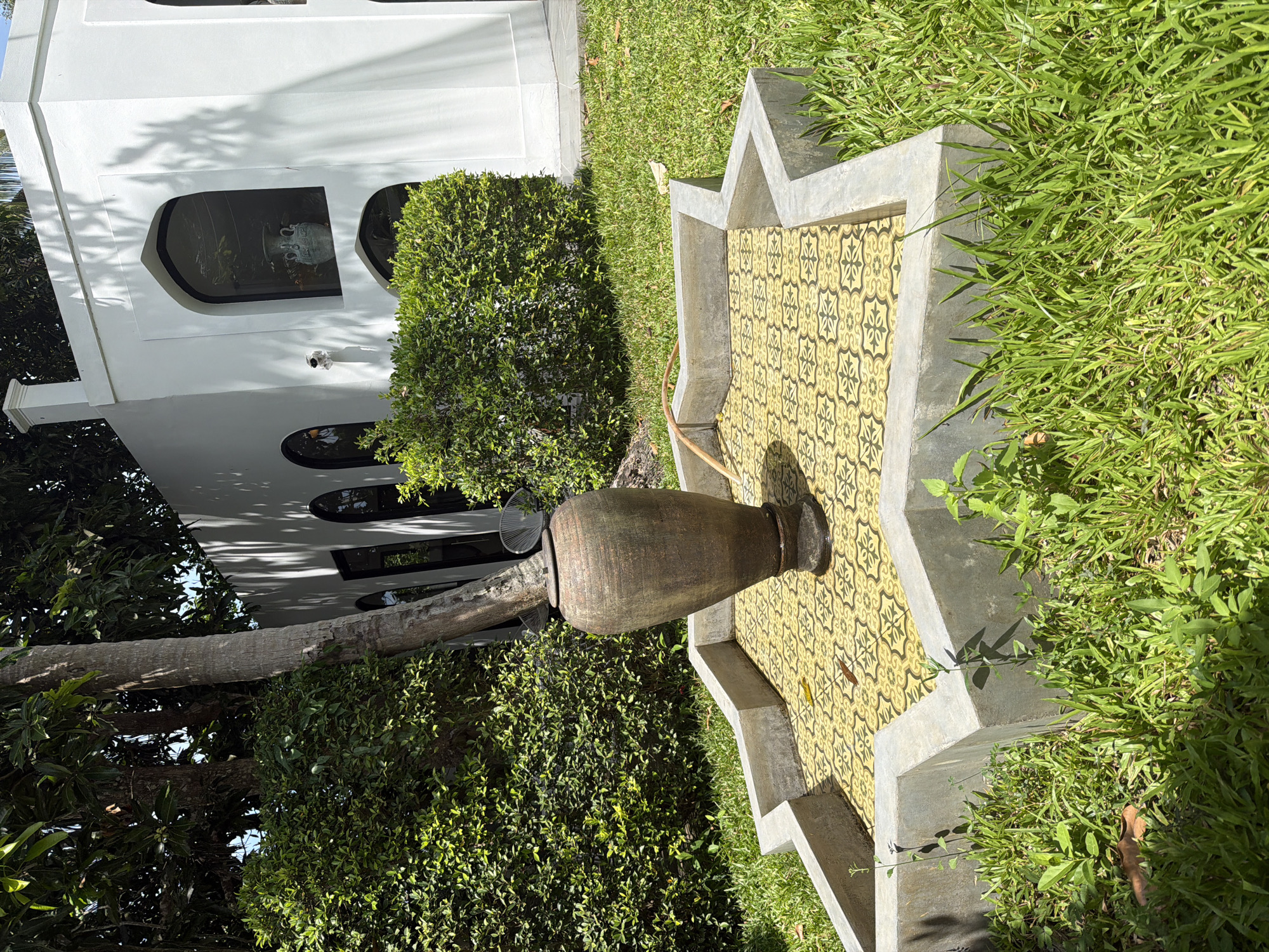 In a sun-drenched outdoor scene, a large, weathered clay pot rests on a patterned yellow tiled surface within a star-shaped concrete planter. The planter is surrounded by vibrant green grass and lush, manicured bushes. In the background, a white building with arched windows and a chimney stands under a canopy of leafy trees, casting shadows on the lawn. The architectural style suggests a resort or a private residence, potentially in a tropical setting like Koh Phangan, Thailand, given the surrounding vegetation and bright sunlight. A thin, light-colored hose snakes across the tiled area, near the base of the pot. No people are visible in the image.