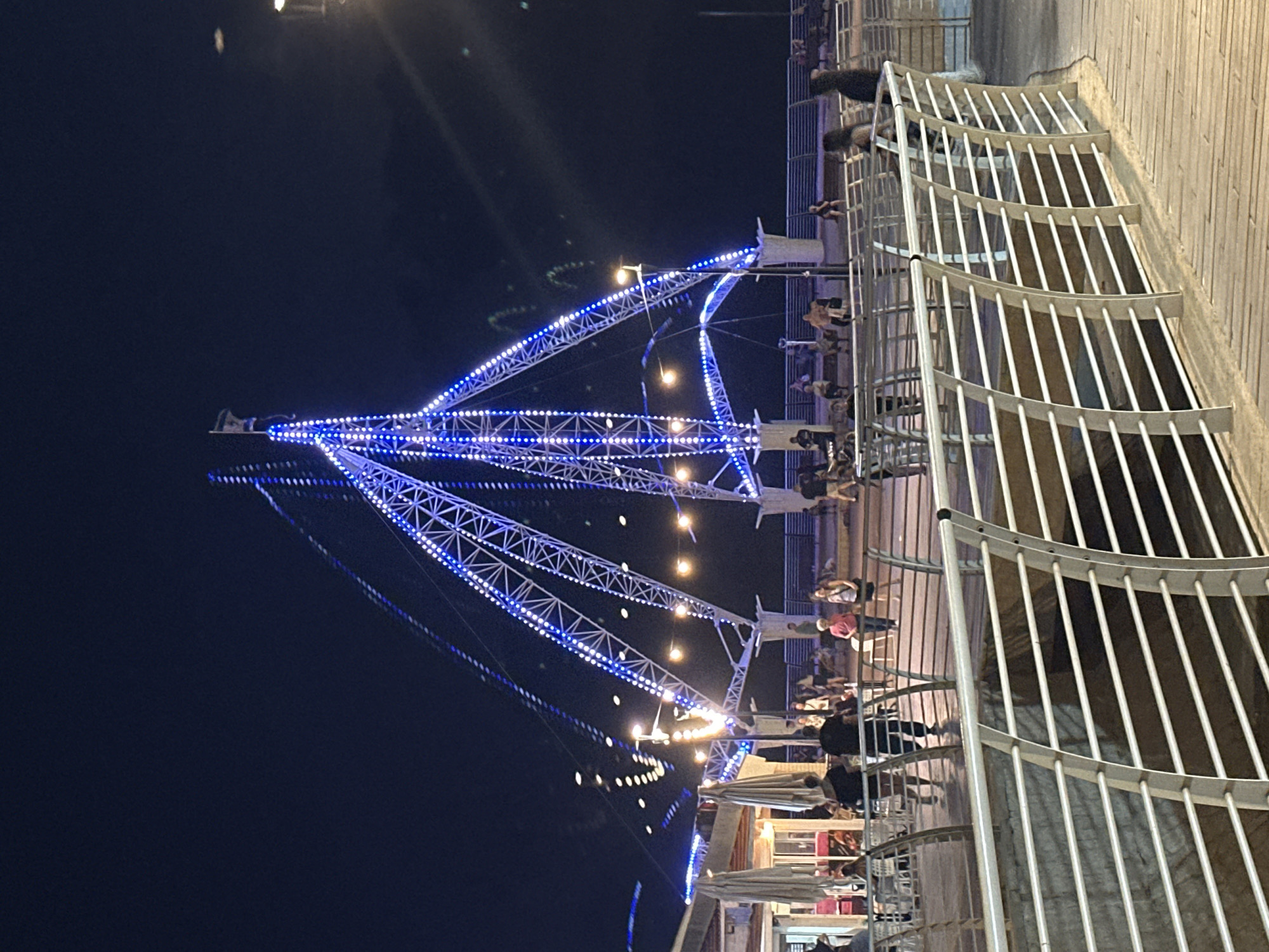 At night in Nahariyya, Israel, a brightly lit Ferris wheel structure with blue lights stands prominently against a dark sky. The structure has a skeletal appearance with triangular supports and is adorned with a dense string of blue LED lights that outline its shape. In the foreground, a wooden boardwalk with a white metal railing curves around the scene. Several people are visible on the boardwalk and in the background, some walking, others seated, and some gathered near a building with outdoor seating and umbrellas. Streetlights with yellow bulbs illuminate parts of the scene, casting light on the walkway and the surrounding area. The overall atmosphere is that of a lively evening at a seaside amusement or recreational area.
