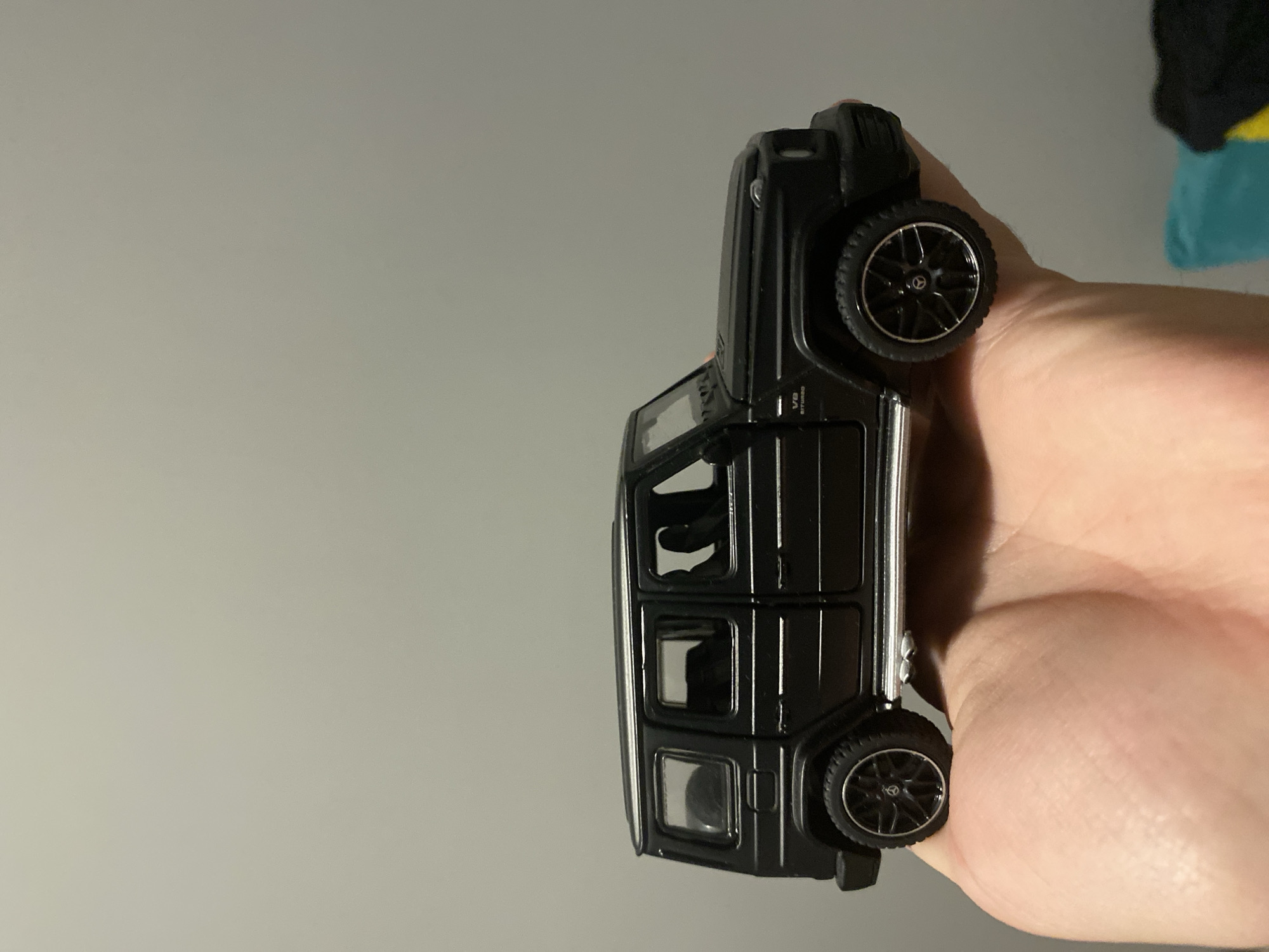 A black Mercedes-Benz G-Class toy car is held in a person's left hand against a plain, grey wall. The toy car is positioned on its side, with the left side facing the viewer. The car appears to be a miniature replica, detailed with realistic features such as the grille, headlights, windows, and wheels. The hand holding the car is large and appears to be an adult's, with visible palm lines and skin texture. The background is a plain, undecorated wall, suggesting an indoor setting. There is no discernible information or context about the location in Łódź, Poland.