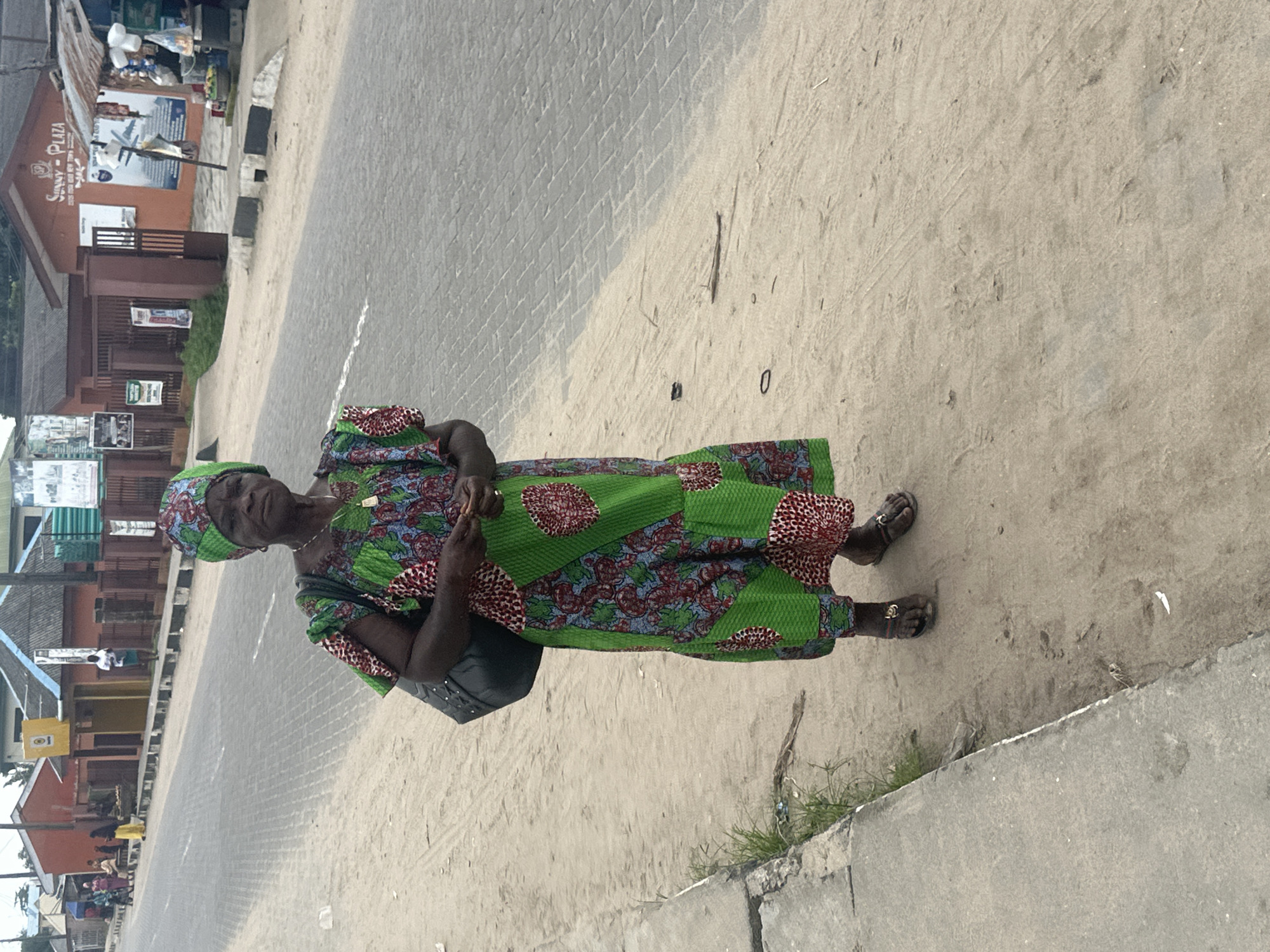 An older Black woman stands on a sandy shoulder next to a paved road in Badagry, Nigeria. She wears a green and red patterned dress and a matching headscarf. A dark shoulder bag is slung across her right shoulder. Her feet are bare, and she wears rings on her toes. She stands with her hands clasped in front of her. In the background, buildings line the opposite side of the road, including one with "Sunny Plaza" signage. A few other individuals are visible in the distance, some near a roadside stall. The road appears to be paved with interlocking bricks.