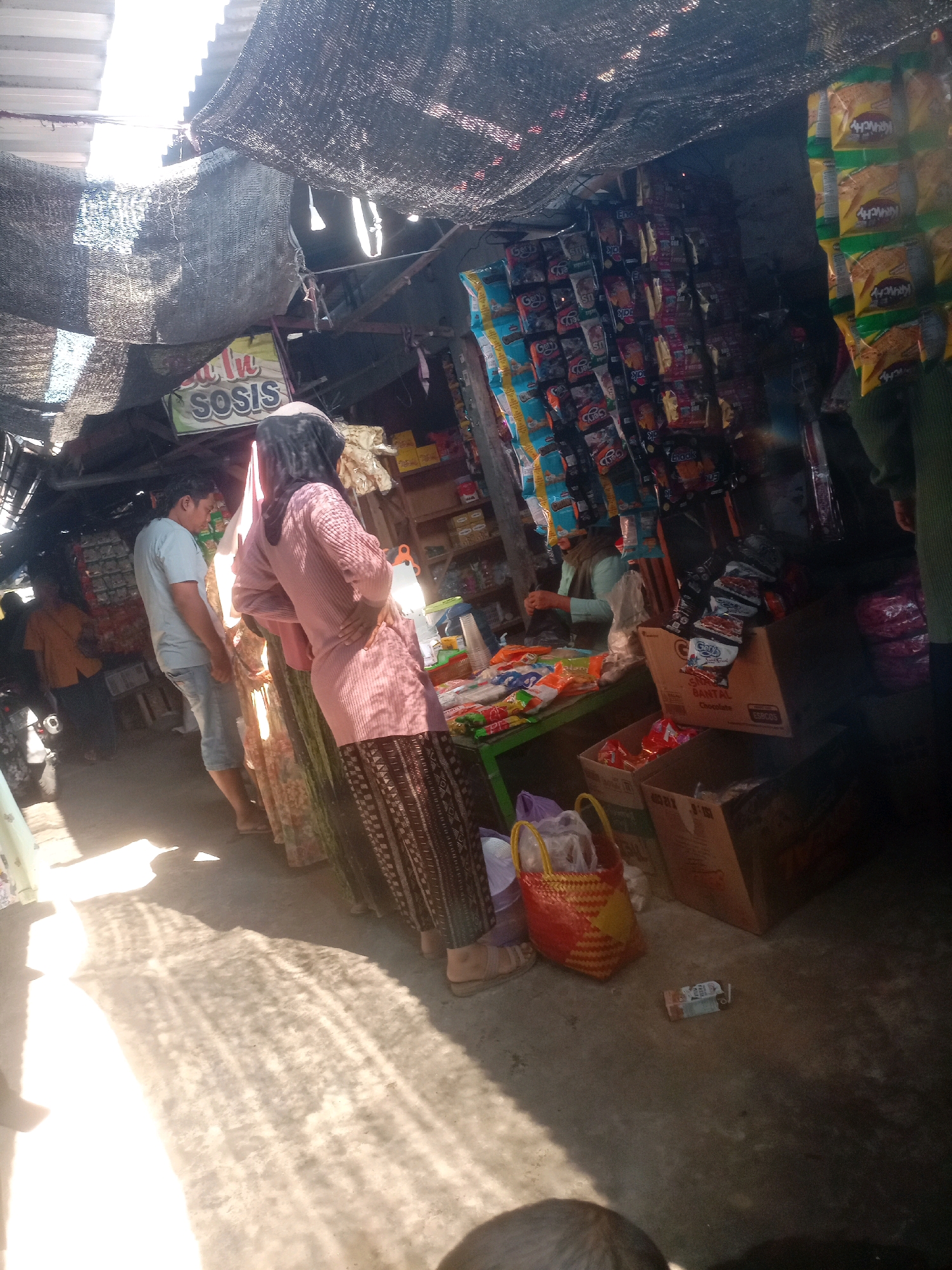 This image depicts a street-side market stall in Sluke, Indonesia, under a corrugated metal roof and dark mesh netting. Several individuals are visible, including a man in a light blue t-shirt and denim shorts, a woman in a pink patterned outfit and headscarf, and another person in an orange shirt standing further back. They appear to be browsing or interacting with the stall's offerings, which include a variety of packaged snacks and other goods displayed on shelves and hanging from the ceiling. A green table is set up in front of the stall, laden with more merchandise, and a woman in a light blue shirt is seen behind it, possibly attending to customers. Cardboard boxes and woven baskets are also present on the ground, containing more items. Sunlight streams in from the left, casting strong shadows on the concrete ground, indicating an outdoor or semi-outdoor setting. The overall atmosphere suggests a busy marketplace environment.