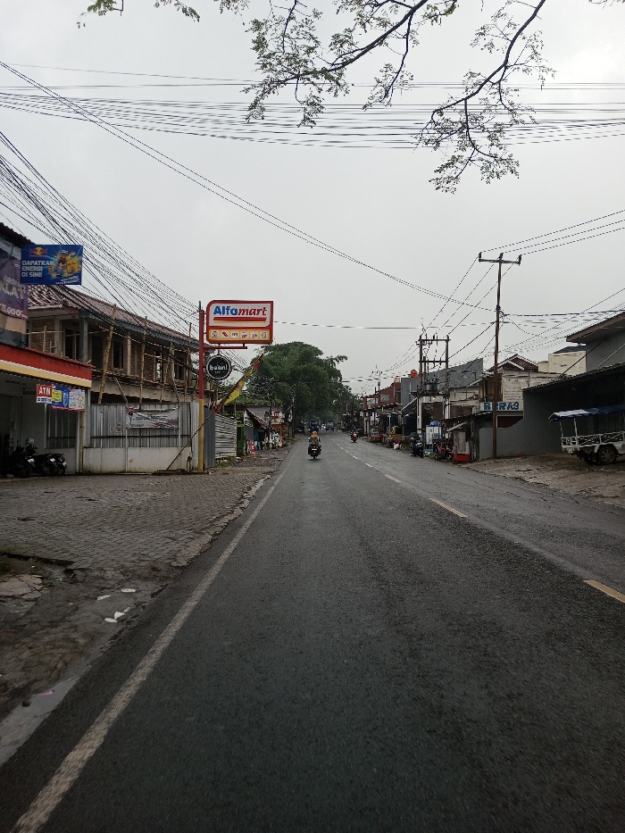 A street view in the city of YonPhoogla, Bhutan, presents a straight asphalt road under an overcast sky. A lone individual on a dark motorcycle travels away from the camera in the right lane; a second motorcycle is visible further in the distance.
On the left side of the road, an uneven paved sidewalk fronts a red-roofed building displaying an ATM sign and a "DAPATKAN DISINI!" banner featuring a Red Bull logo. Immediately adjacent is a multi-story building under construction, characterized by exposed bamboo scaffolding and a grey corrugated metal fence at ground level. Further down the street, a prominent "Alfamart" sign is mounted on a pole, with a smaller circular "baani" sign positioned below it. Multiple motorcycles are parked on the left sidewalk.
The right side of the road features a continuous line of commercial and residential structures, including one building with a visible "BERAS" sign. A light-colored pickup truck with a blue tarp covering its bed is parked on the far right. Numerous utility poles line both sides of the street, supporting a dense network of overhead electrical and communication wires that crisscross the entire scene. Bare tree branches extend into the upper left portion of the frame against the bright, cloudy sky.