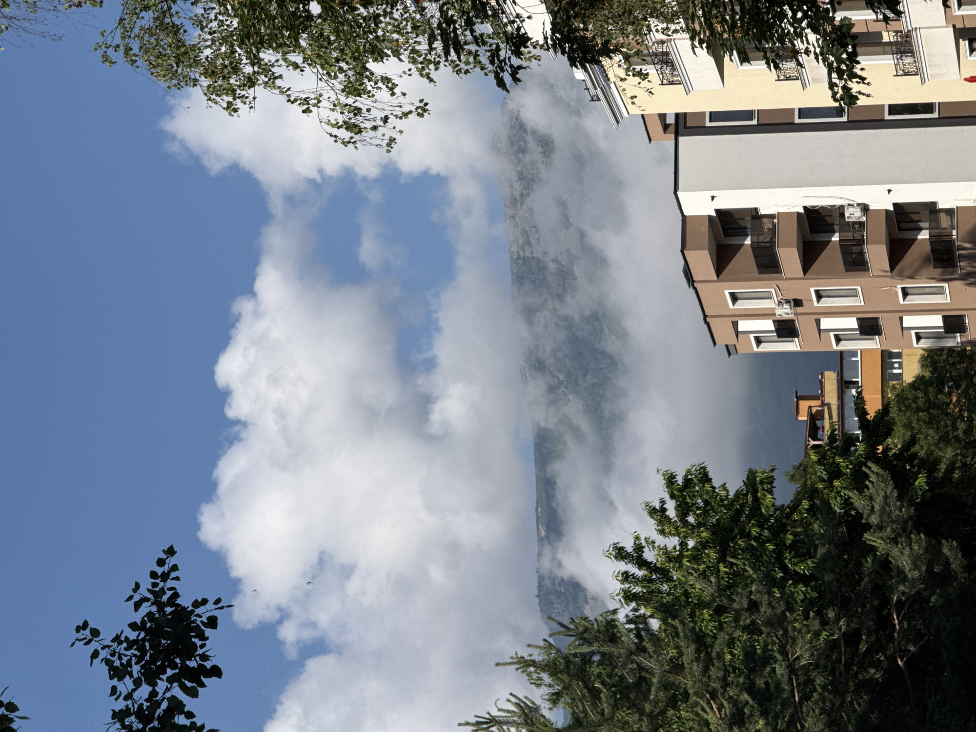 This image captures a vibrant urban and natural landscape in Sofia, Bulgaria, depicting multi-story buildings set against a backdrop of majestic, cloud-shrouded mountains under a bright sky.
In the upper part of the frame, a clear blue sky is visible, adorned with scattered, fluffy white cumulus clouds. Below this, a large mountain range stretches across the mid-ground, its peaks and slopes partially obscured by a thick layer of white mist or low-hanging clouds, creating an ethereal and dramatic effect.
The foreground and mid-ground feature several contemporary residential or apartment buildings. On the right, a building with light brown and white facades, multiple windows, and balconies with black railings is prominent. Below it, a larger, brown-colored building with distinct white accents and recessed rectangular windows stands out, with an air conditioning unit visible on one of its walls. Further towards the left and lower, a yellow-orange building is partially visible, peeking through the trees.
Lush green trees are interspersed throughout the scene. In the upper left, branches with vibrant light green leaves extend into the sky. The lower and left sections of the image are filled with a dense canopy of darker green foliage, comprising both broadleaf and coniferous trees, framing the urban structures and mountain view.
The photograph depicts a bright, clear day, likely late morning or early afternoon, as evidenced by the strong natural light and visible shadows on the buildings. The weather appears partly cloudy with localized mist around the mountains. The scene emphasizes the harmonious coexistence of modern city architecture with the imposing natural beauty of nearby mountains. No people or visible text are present in the image.