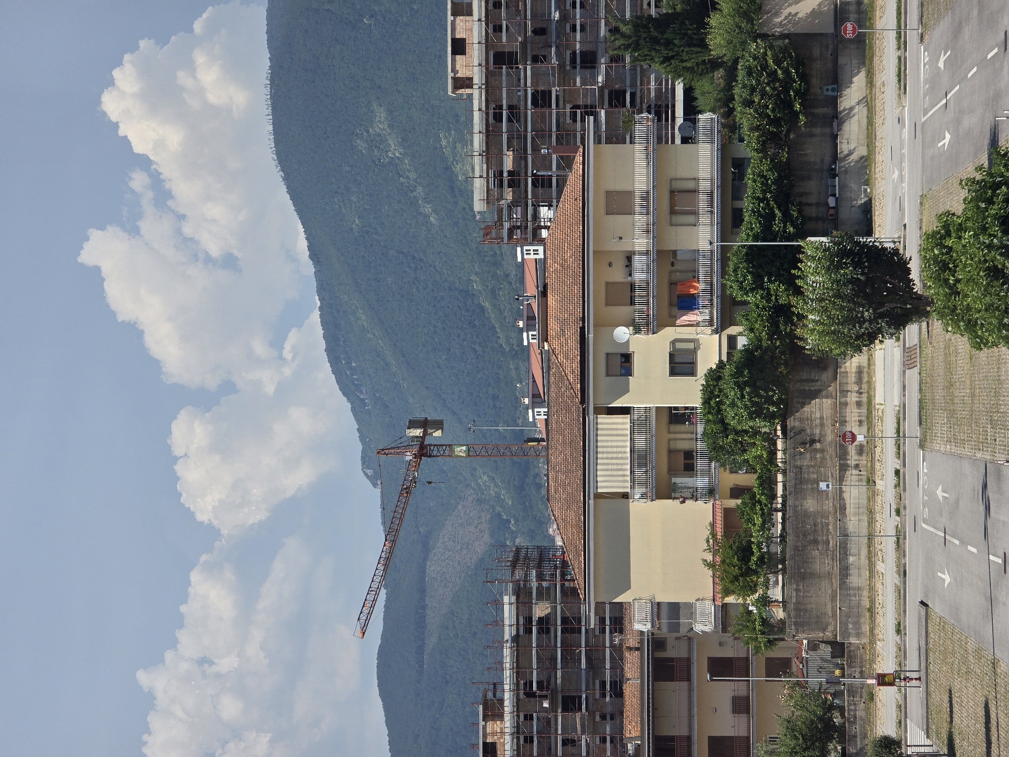 The image depicts an urban scene in Baronissi, Italy, dominated by construction and residential buildings set against a backdrop of lush green mountains under a partly cloudy sky. In the foreground, a paved road with white directional arrows and "STOP" markings suggests an intersection or parking area. A red fire hydrant and a lamppost stand on the left side of the road.
Behind the road, a concrete retaining wall separates it from a strip of greenery and several trees. Beyond this, a pale yellow multi-story residential building with balconies features awnings and laundry hanging out to dry. Adjacent to it, a partially constructed building is visible, encased in scaffolding, with exposed brickwork and window openings.
A prominent orange construction crane stands tall to the left of the unfinished building, its boom extending diagonally across the frame. Further in the background, more multi-story buildings, also appearing to be under construction or renovation, are present. The mountains in the distance are covered in dense vegetation, with visible patches of cleared land on their slopes. The overall impression is one of ongoing development within a scenic, natural setting.